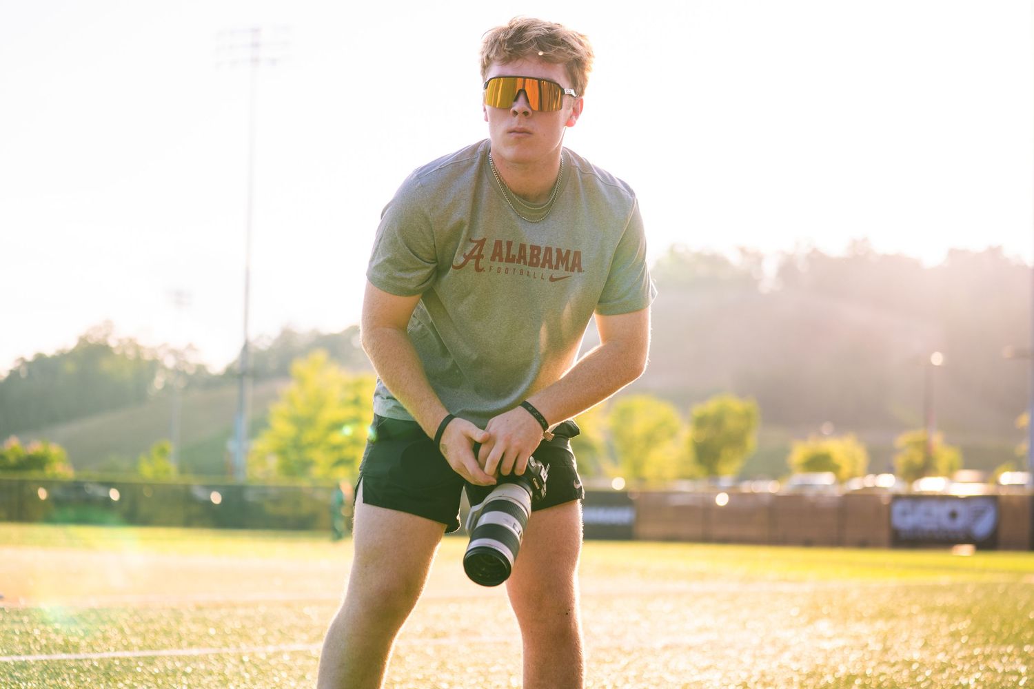 Photographer in green shirt and shorts adjusts camera equipment on sunny sports field at golden hour.