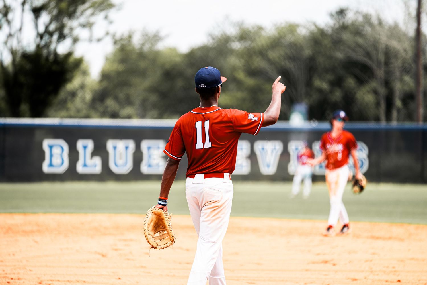Baseball player wearing jersey number 11 raises hand on field in front of Blue Devils sign.