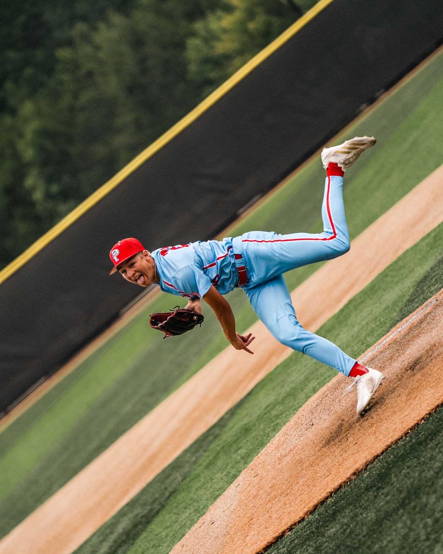 Baseball pitcher in light blue uniform and red cap mid-throw on the mound during a game.