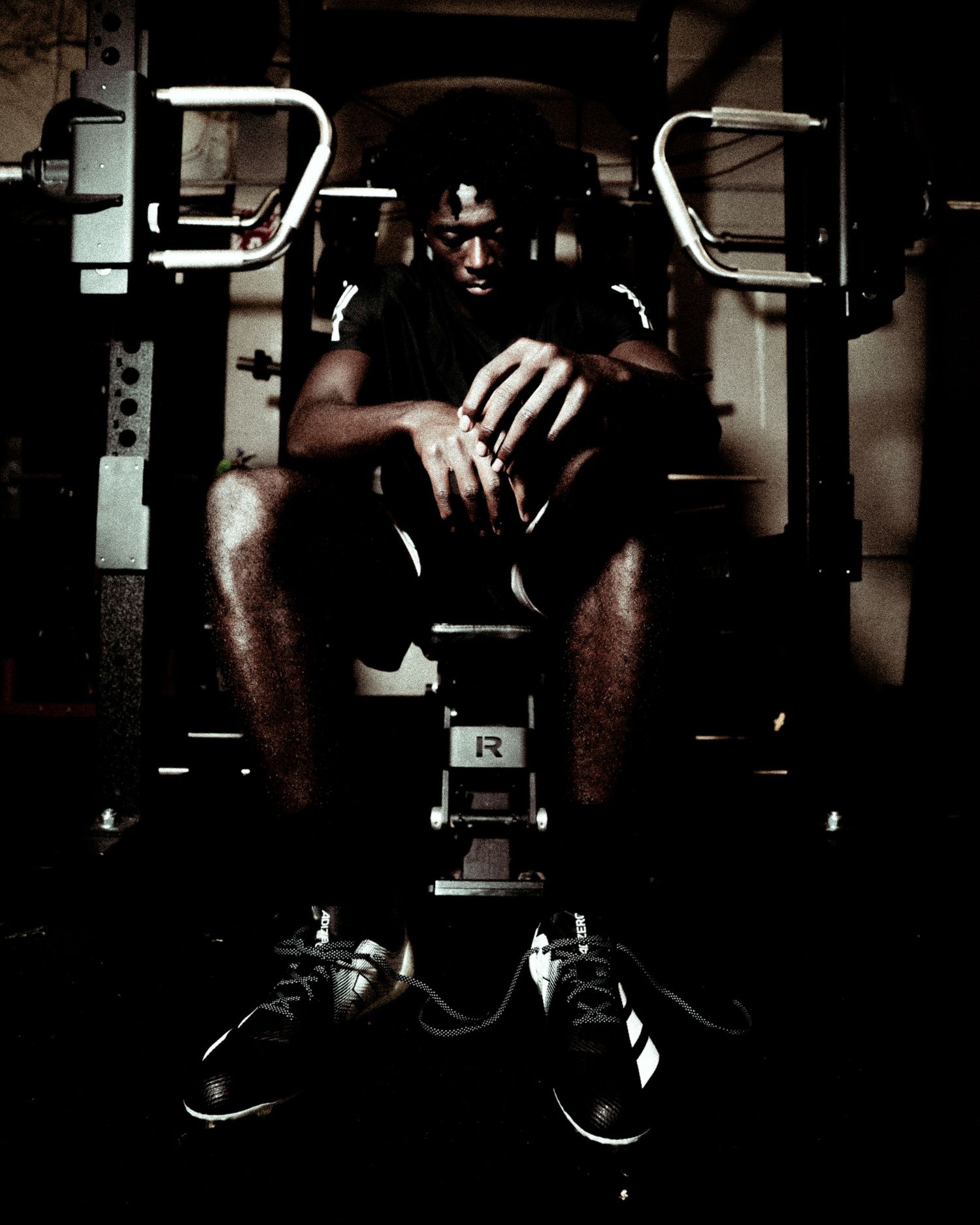 Athlete sits in gym equipment in dramatic low-key lighting, wearing black shorts and Adidas shoes.