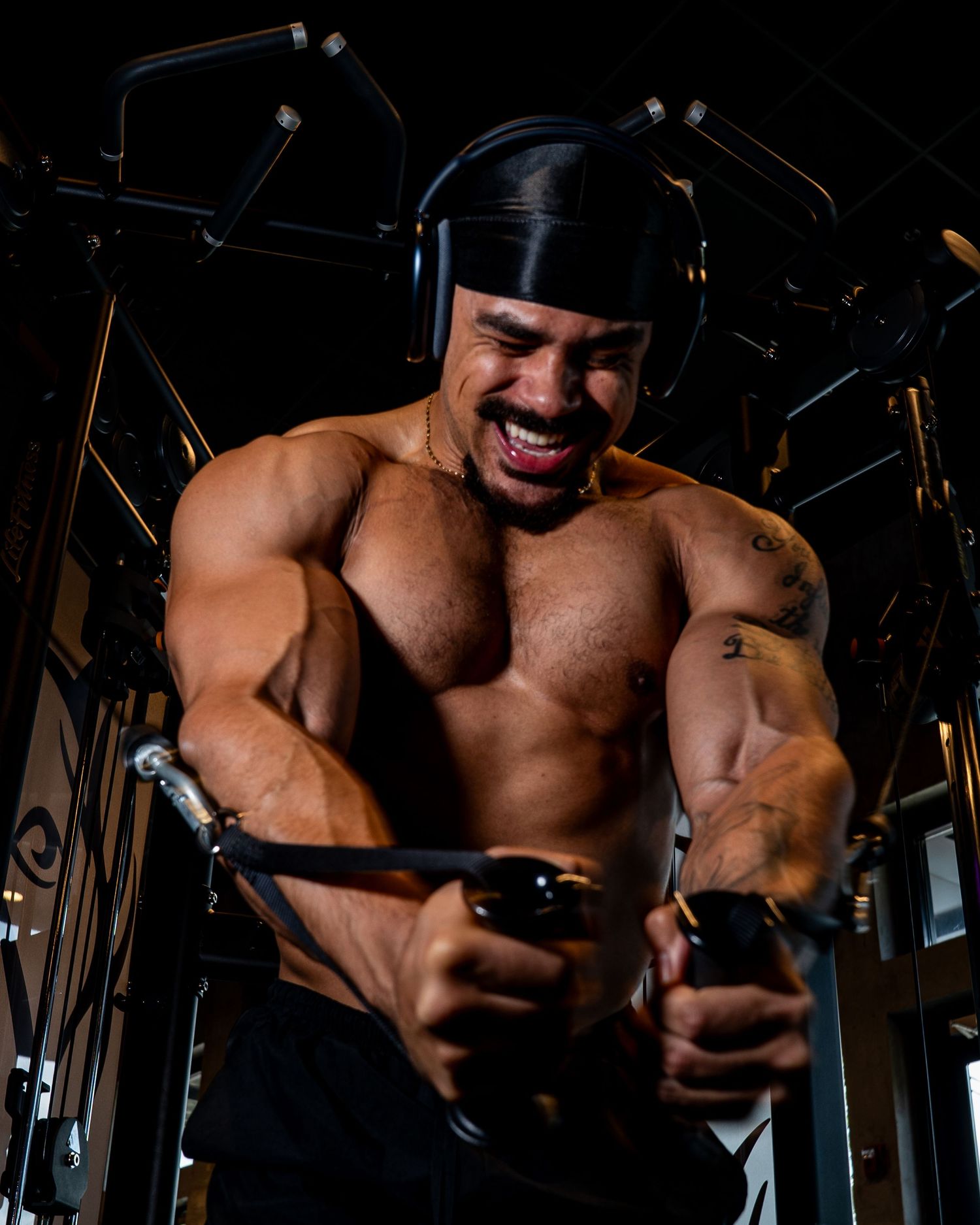 Muscular bodybuilder intensely performing cable fly exercise in gym, showcasing massive chest and arm muscles while grimacing.