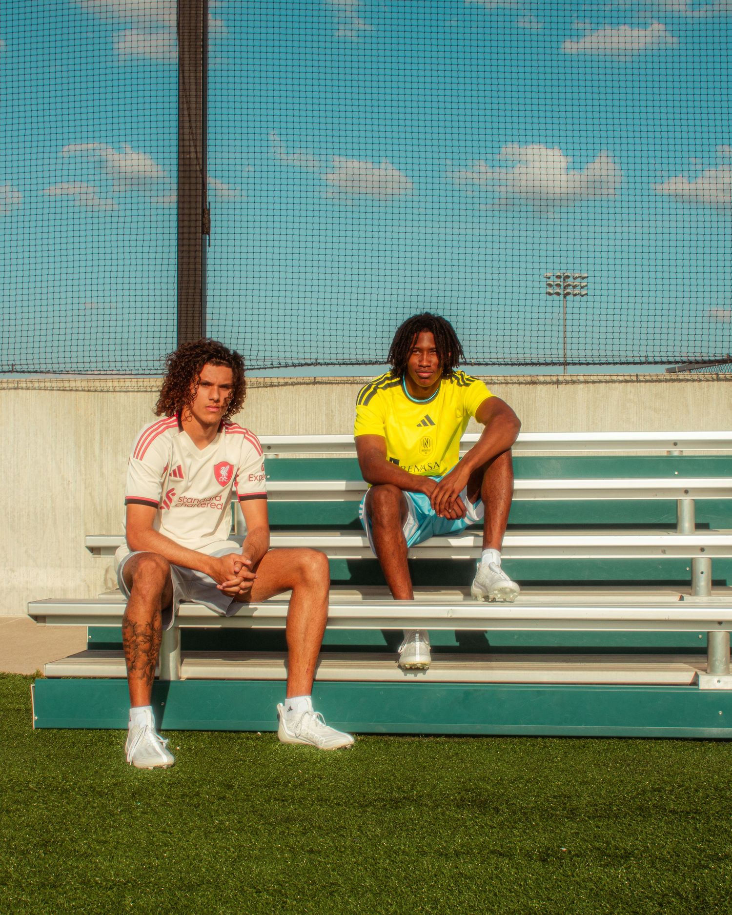 Two soccer players in Adidas jerseys sit on bleachers at a sports field under a blue sky.