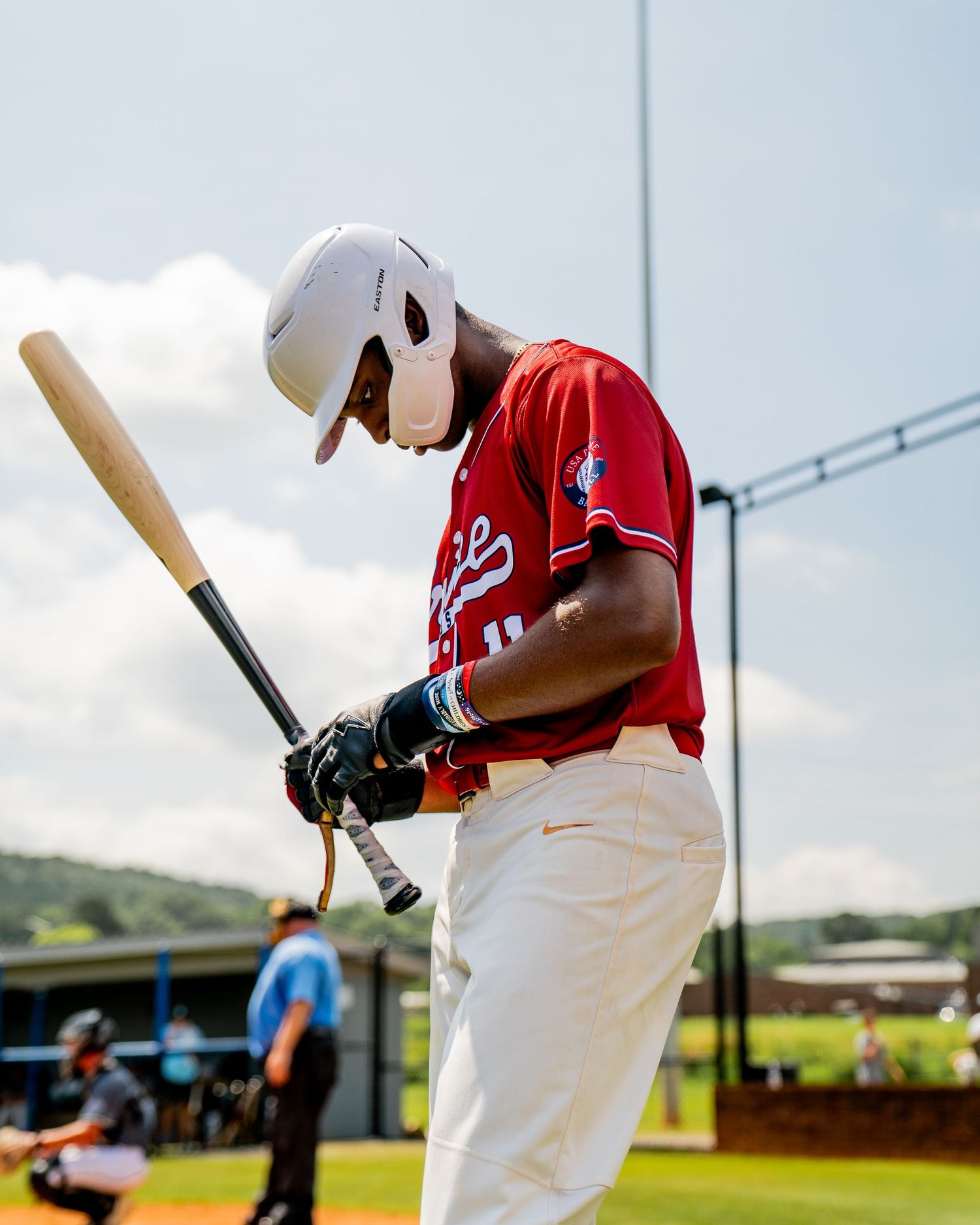 A baseball player in a red jersey and white helmet holds a bat while looking down on a sunny day at the ballpark.