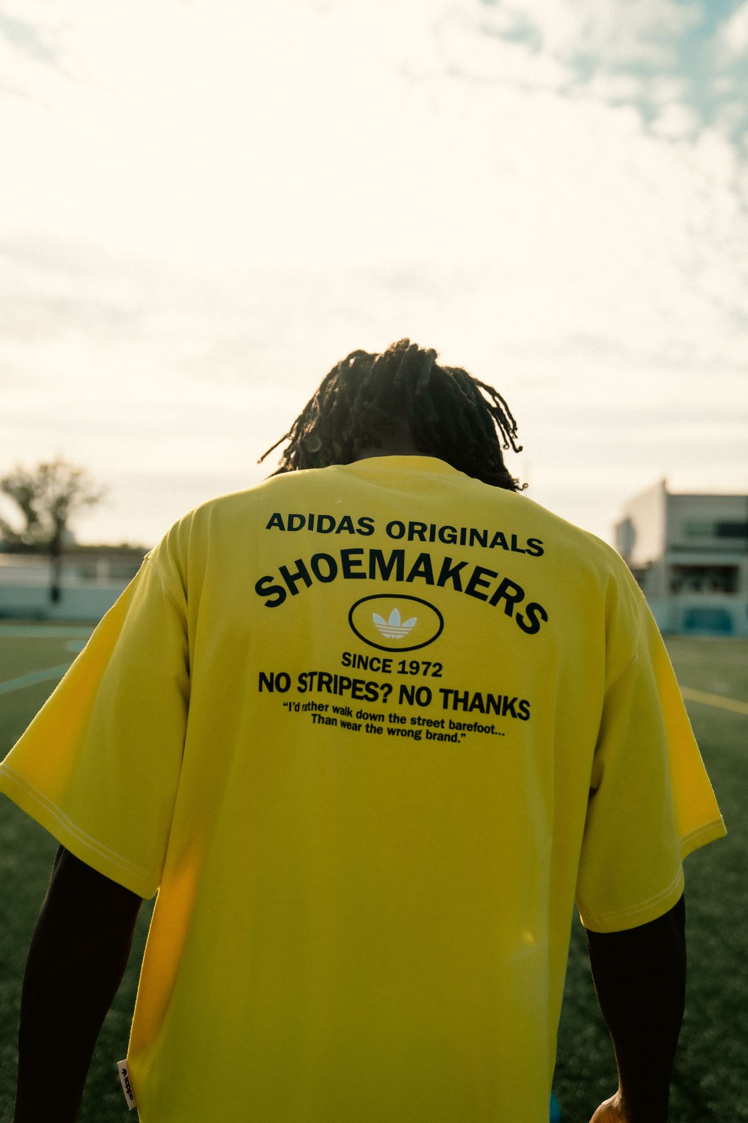 Person wearing yellow Adidas Originals Shoemakers graphic t-shirt on a sports field.