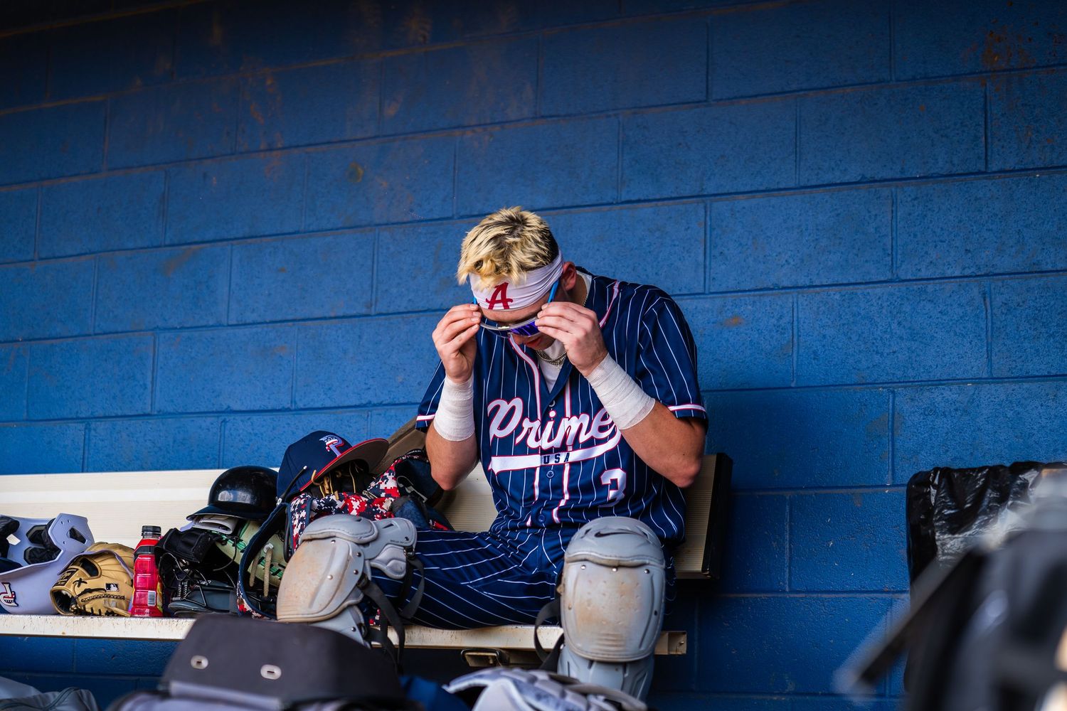 Baseball player in Prime jersey sits in dugout adjusting sunglasses, surrounded by gear against blue wall.
