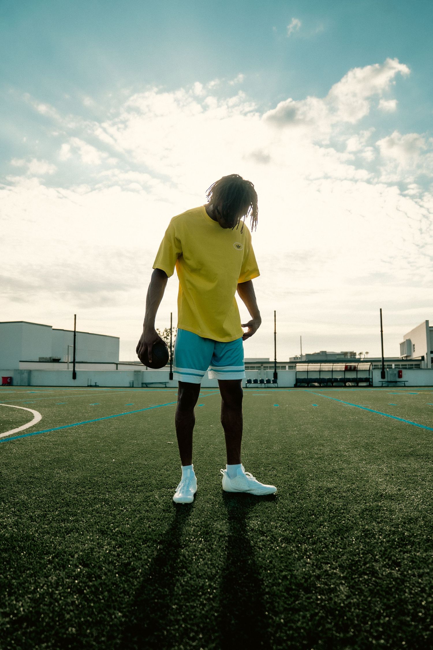 Young man in yellow shirt and blue shorts holding a basketball on an outdoor court under a cloudy sky.