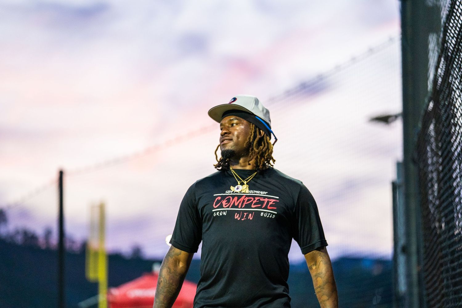Man with dreadlocks wearing a Compete brand shirt and cap stands outdoors at dusk with a colorful sky.