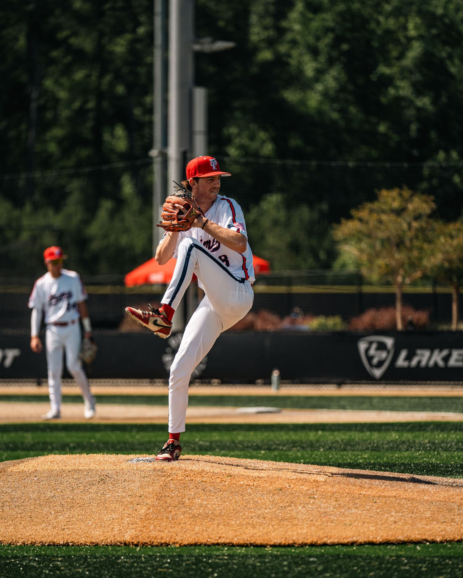 Baseball pitcher in white uniform winds up to throw on a sunny outdoor mound with green trees in background.