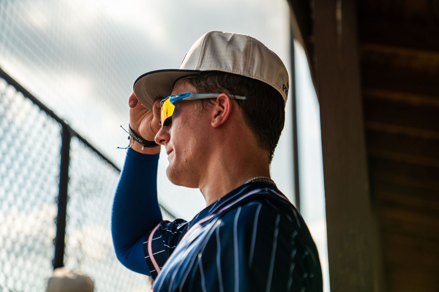 A young baseball player in a blue pinstripe jersey and white cap adjusts his yellow sunglasses in a dugout.