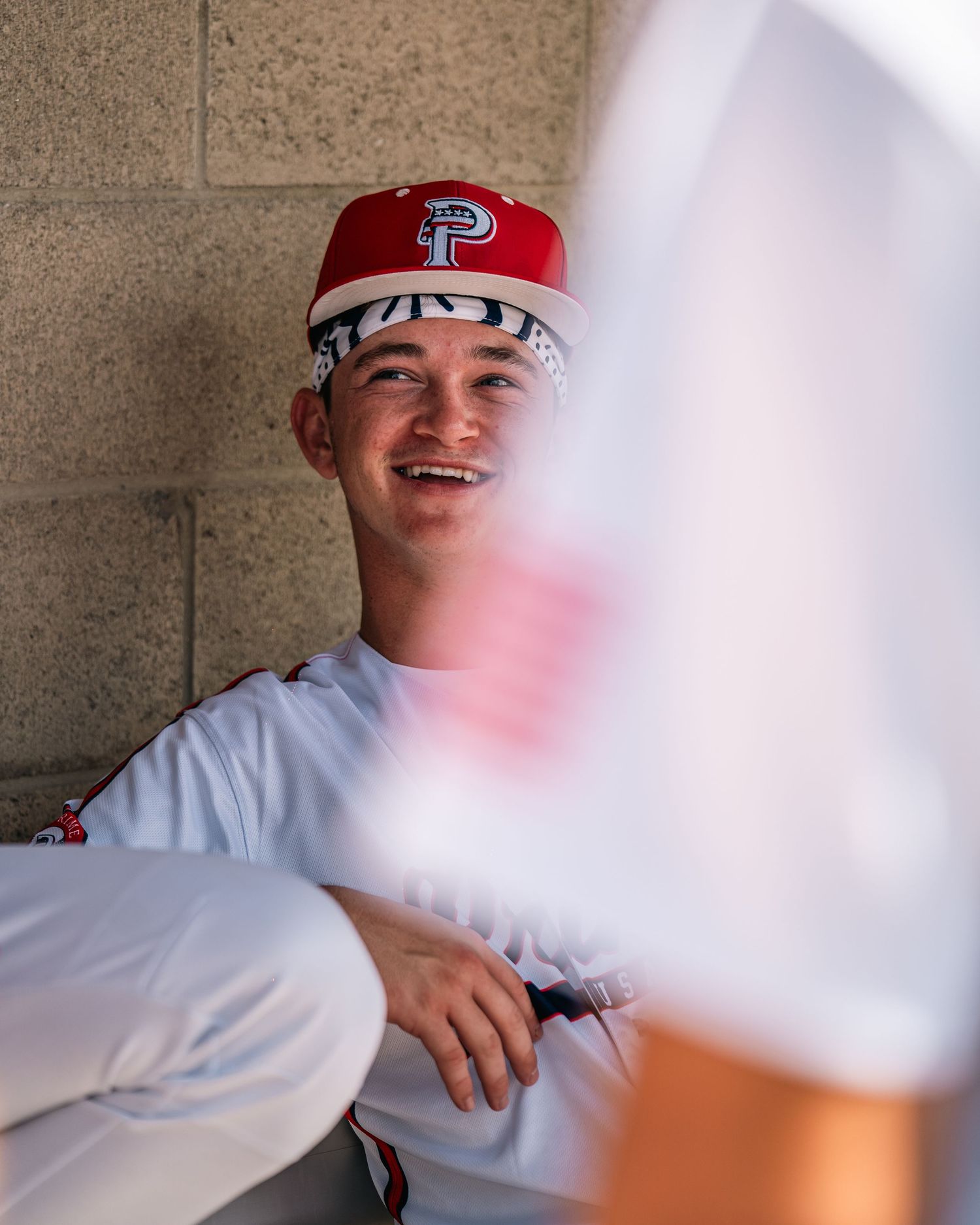 Smiling baseball player in dugout wearing red Prime cap and white uniform.