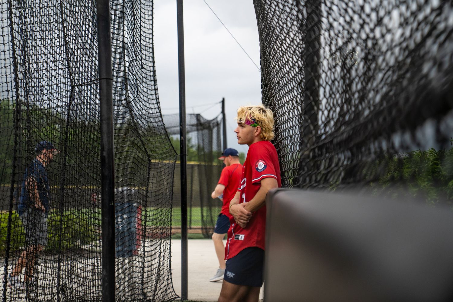 Young baseball players in red uniforms stand near batting cage nets during outdoor practice session.