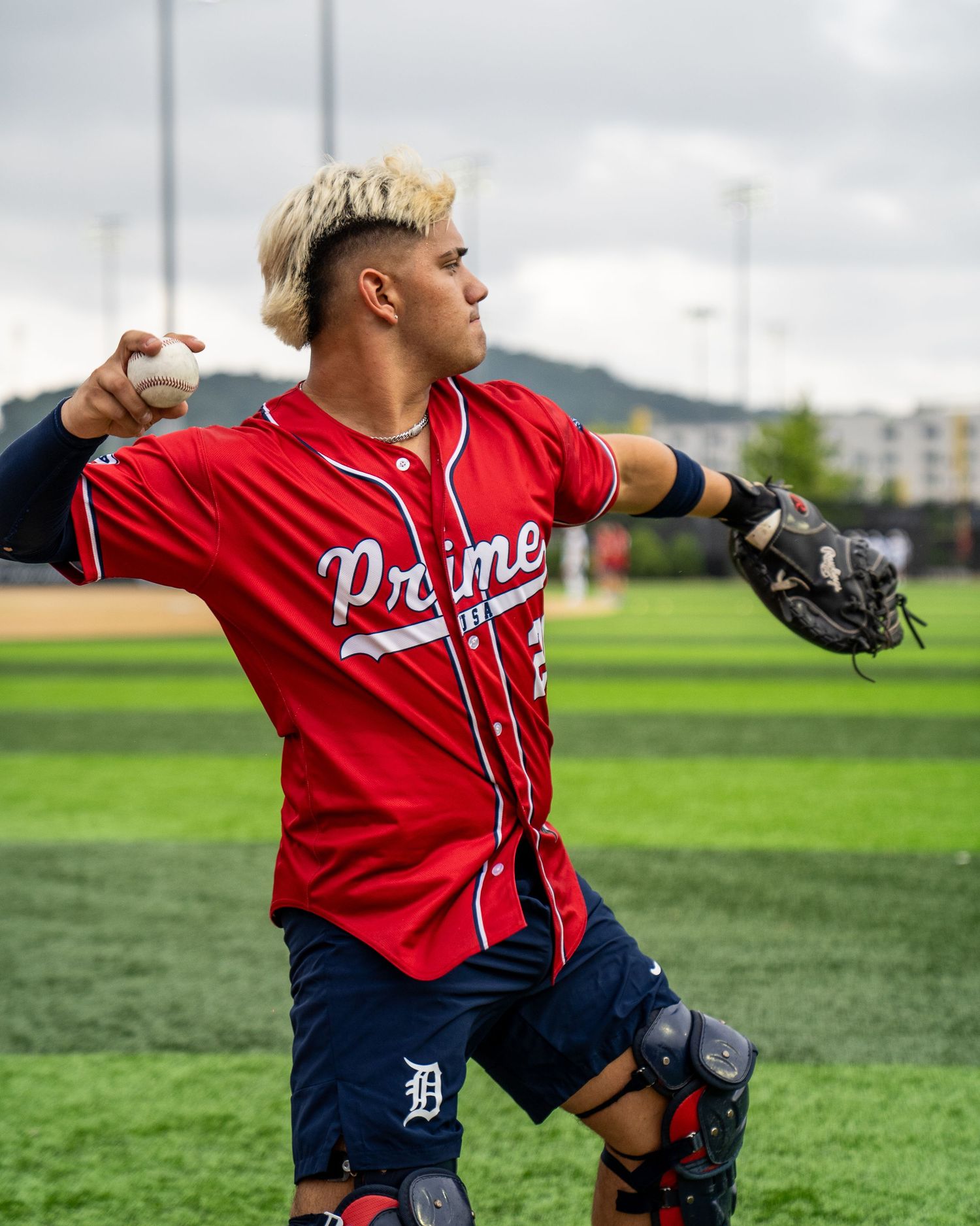Baseball player in red Prime jersey throwing ball on green field wearing catcher gear and Detroit Tigers logo shorts.