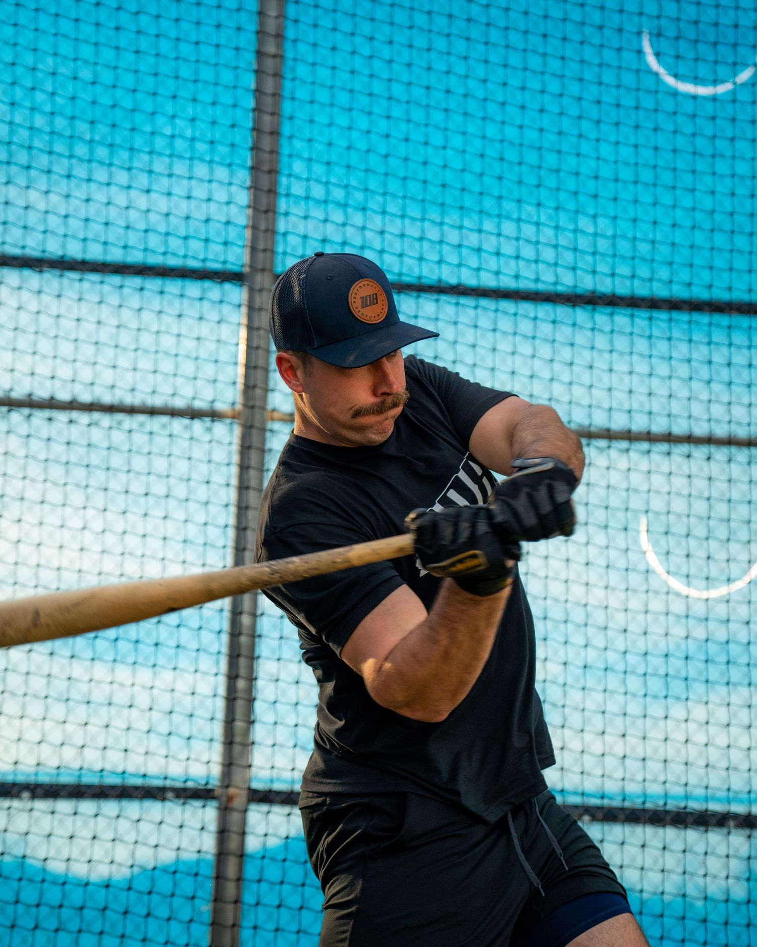 Man in black outfit swinging a baseball bat in batting cage wearing navy snapback cap and batting gloves.