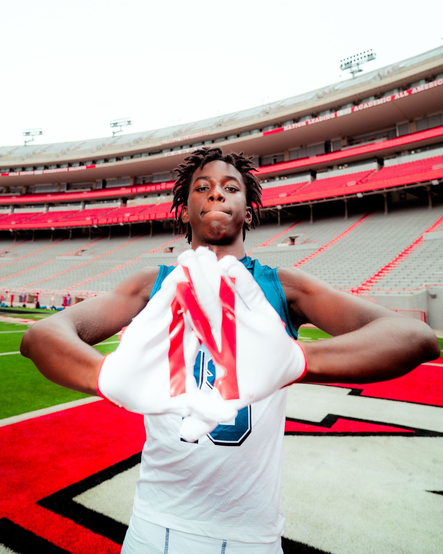 Young athlete poses in stadium holding jersey number, standing on field with red track and bleachers in background.