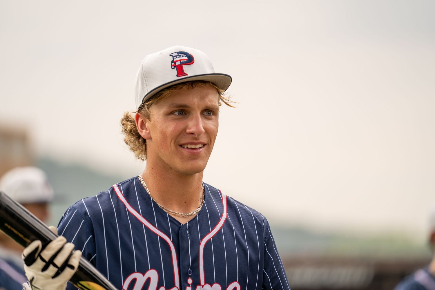 Smiling baseball player in navy pinstripe uniform and white cap holding a bat outdoors.