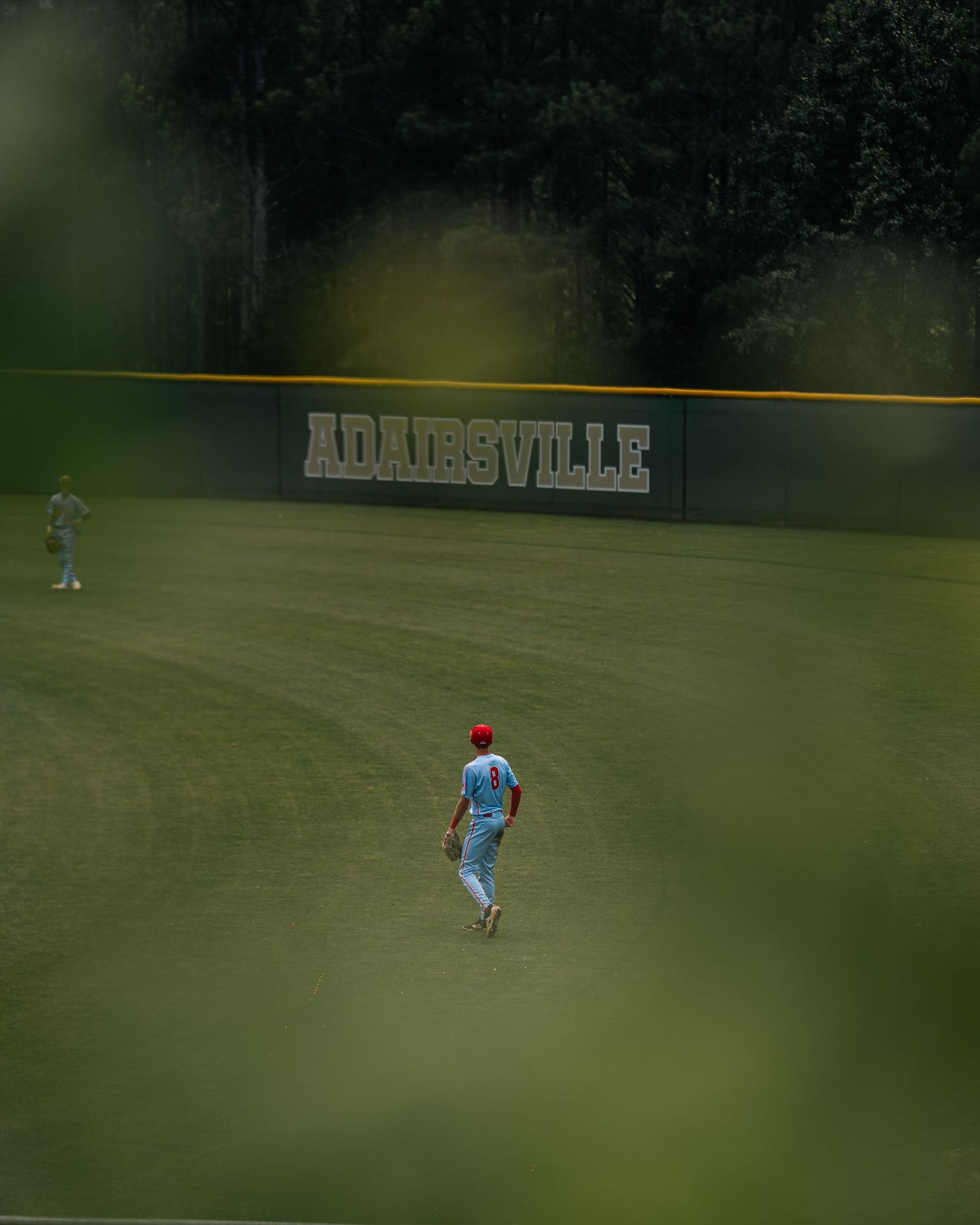 Baseball players on the field at Adairsville stadium, surrounded by lush green grass and dense trees.