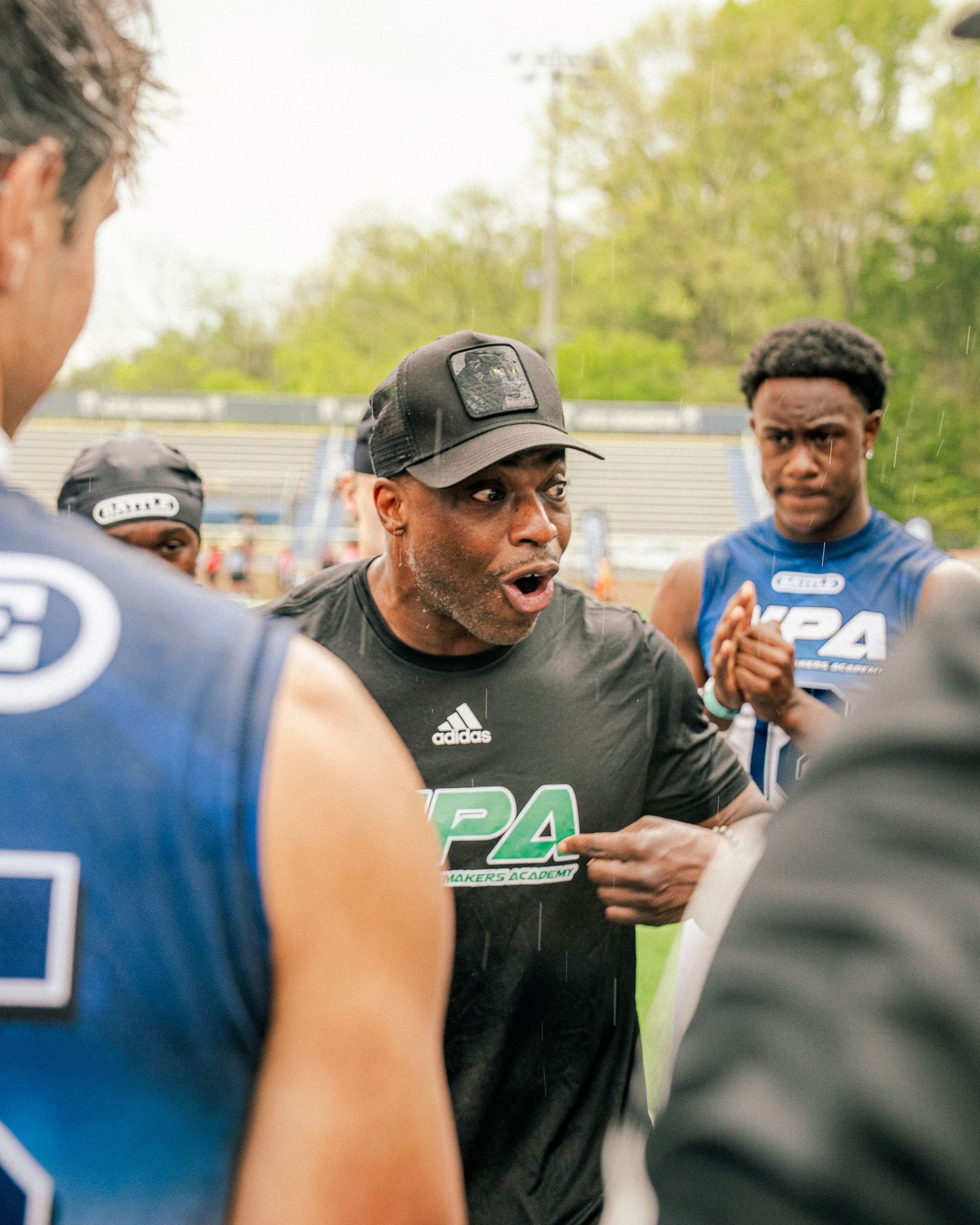 Coach in NPA shirt motivates players in huddle at outdoor football training event.