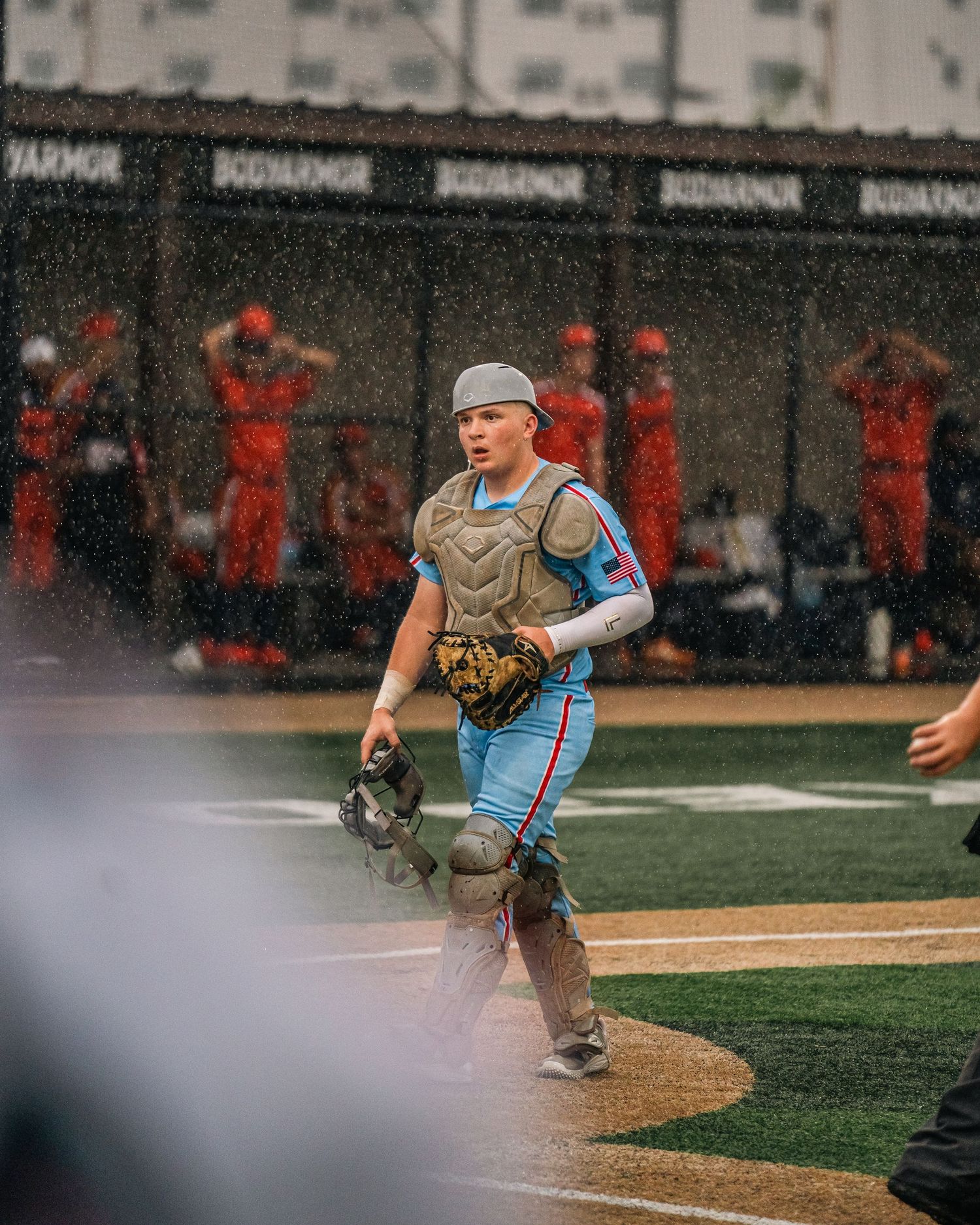 Baseball catcher in blue uniform and chest protector stands alert on rainy field during game.