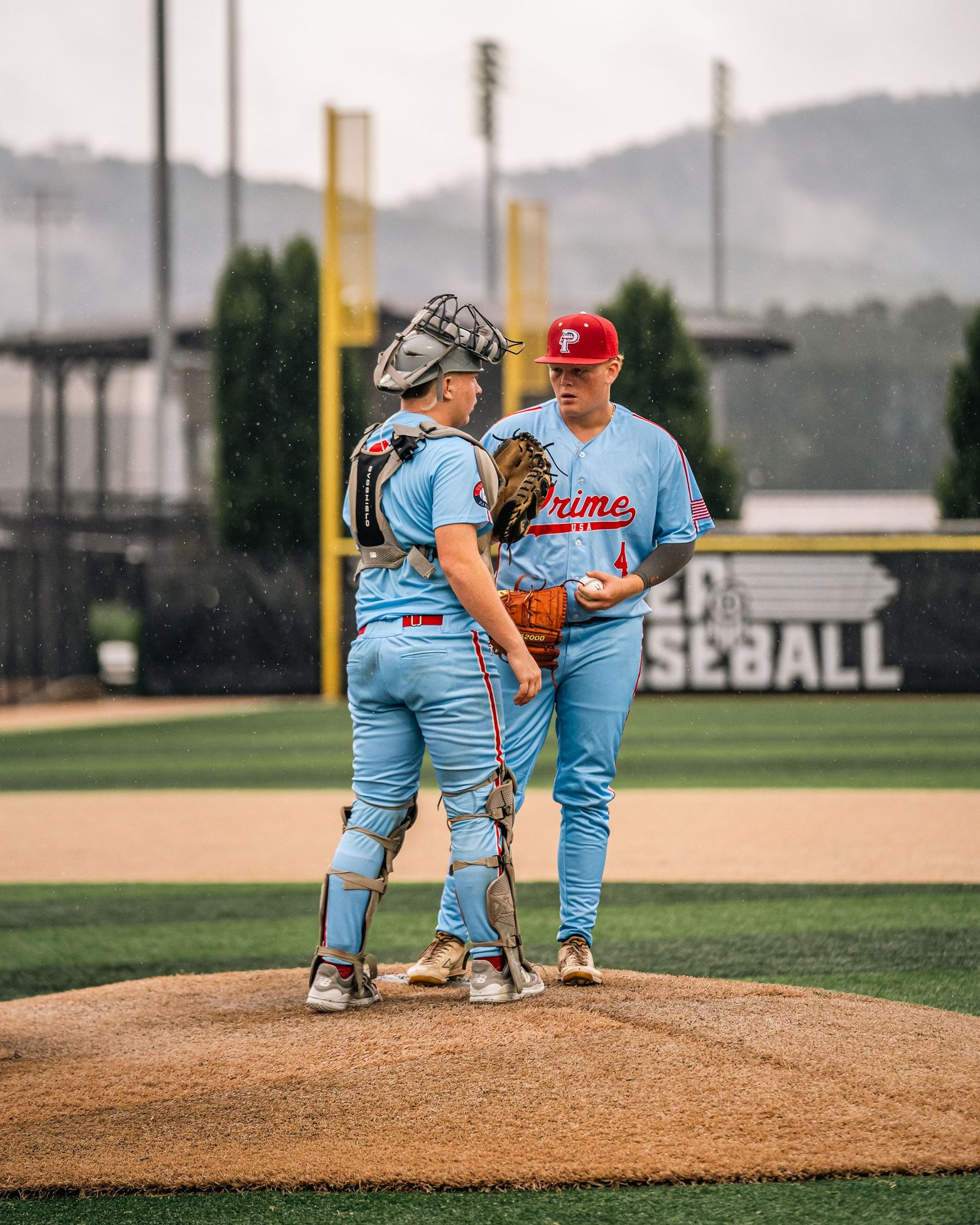 Baseball catcher and pitcher in light blue Prime uniforms conferring on the mound.