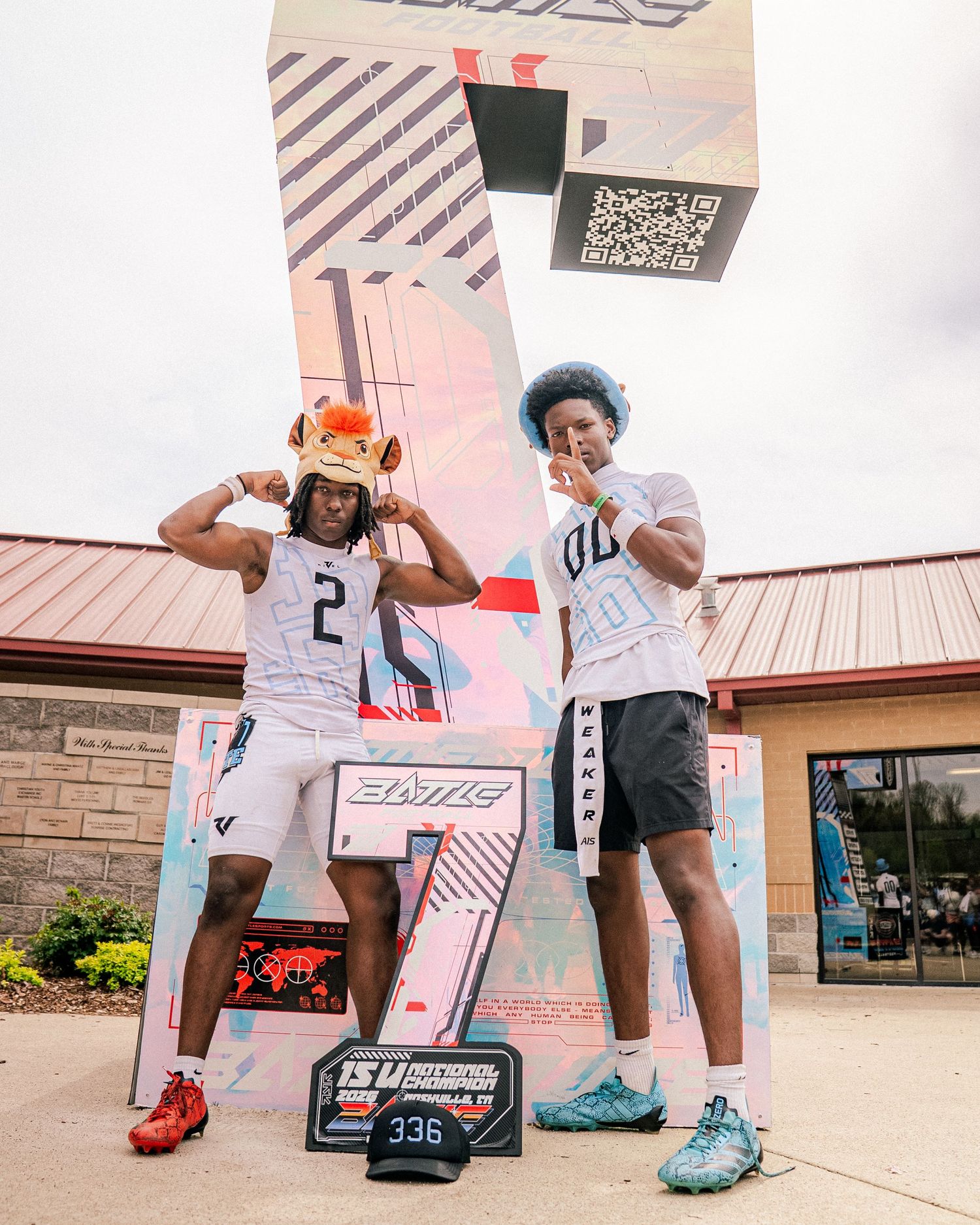 Two football players in white uniforms pose confidently in front of a colorful sports event display outdoors.