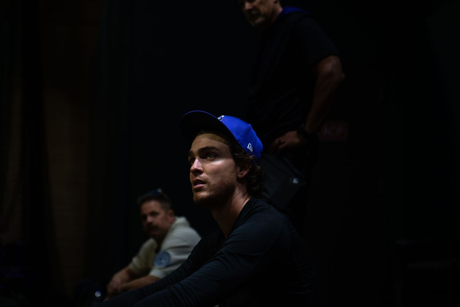 Young man wearing a blue snapback cap and dark shirt sitting in a dimly lit room.