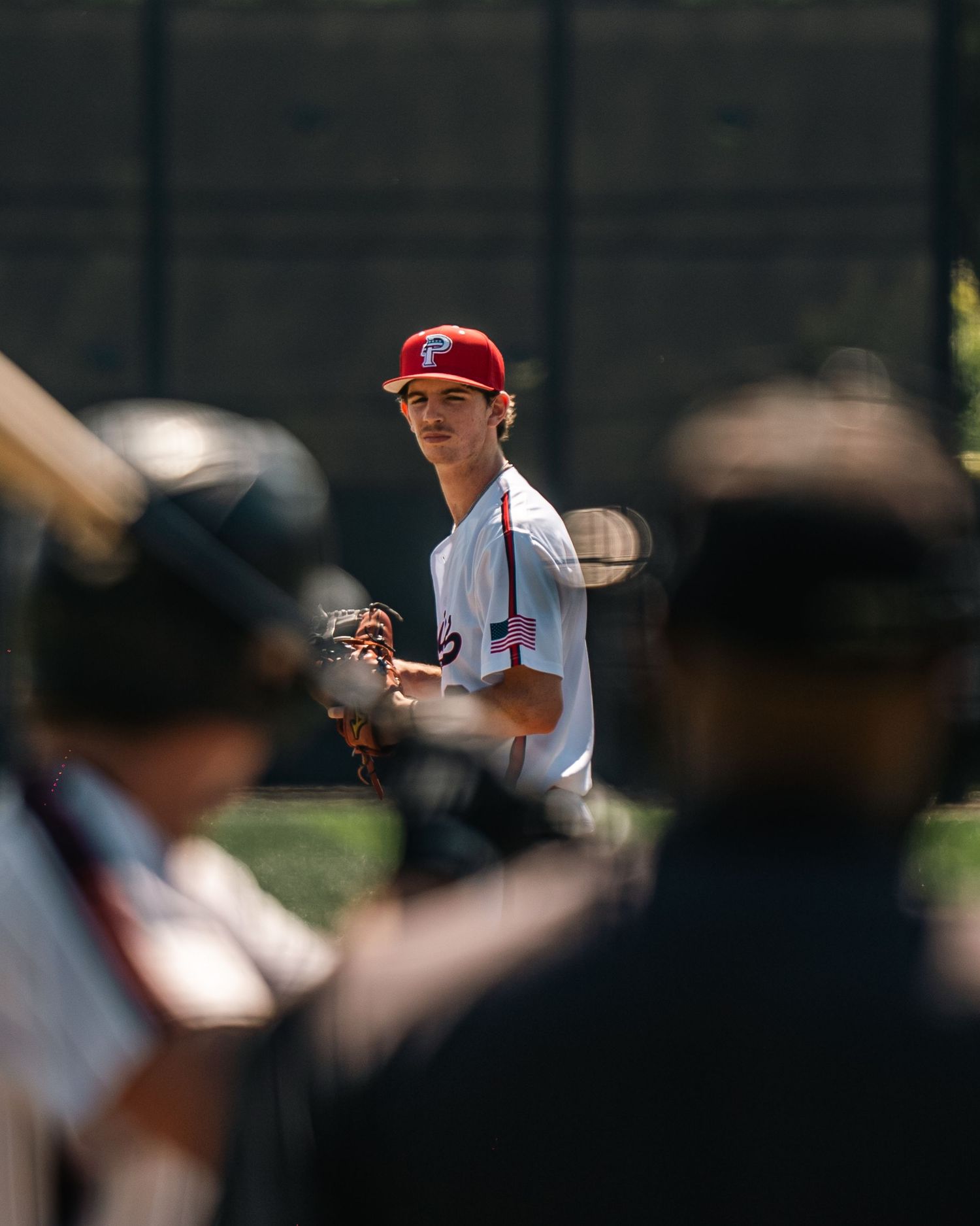 A young baseball pitcher in a red cap and white uniform stands ready on the mound during a sunny game.