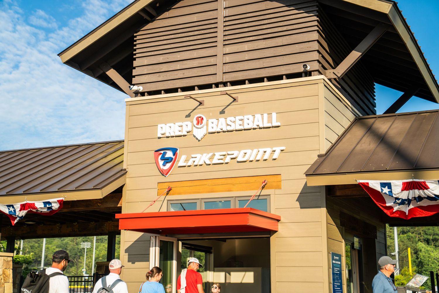 Prep Baseball Lakepoint facility entrance with signage, patriotic banners, and visitors outside on a sunny day.