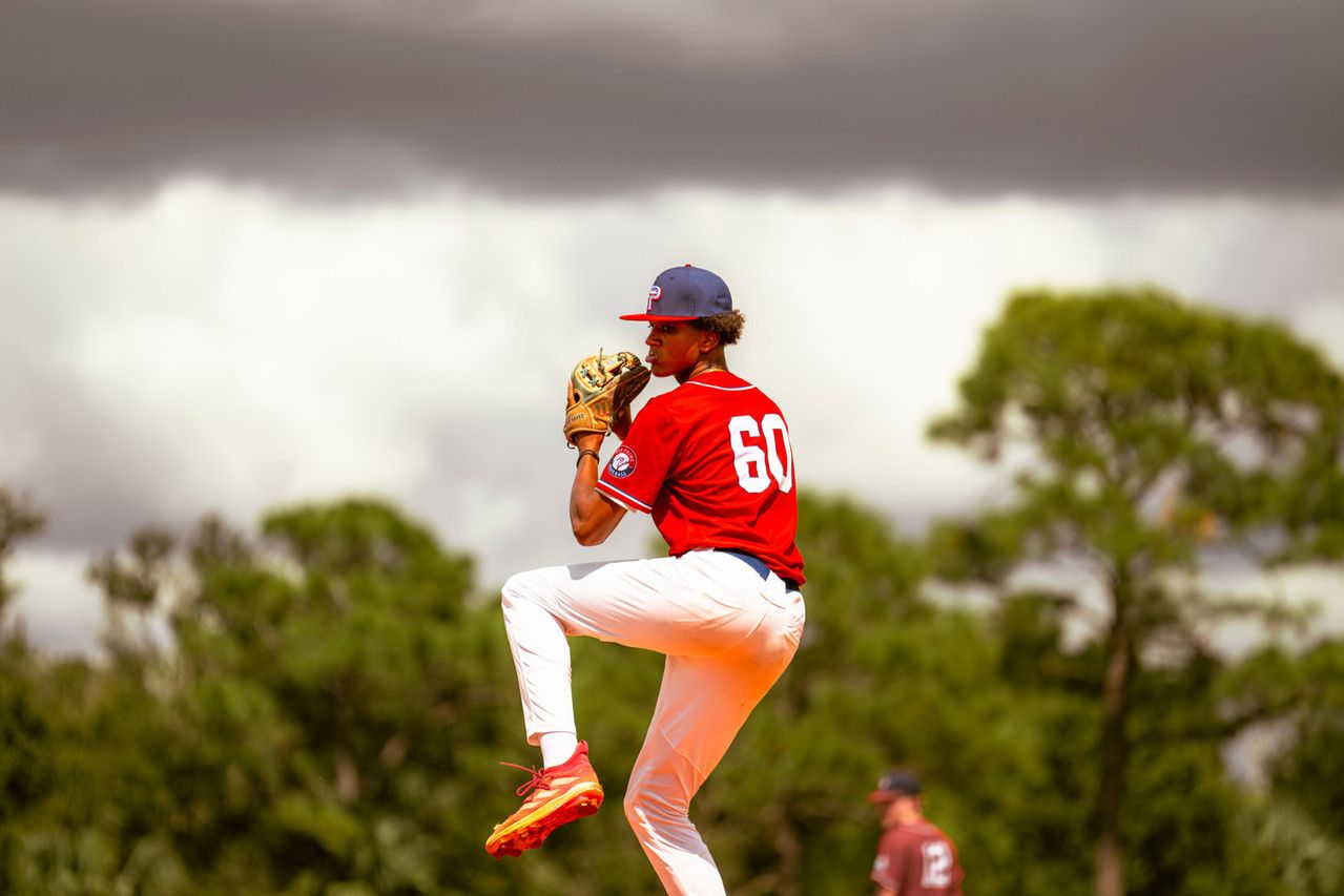 Baseball pitcher in red uniform, number 60, mid-windup on mound under dramatic cloudy sky.