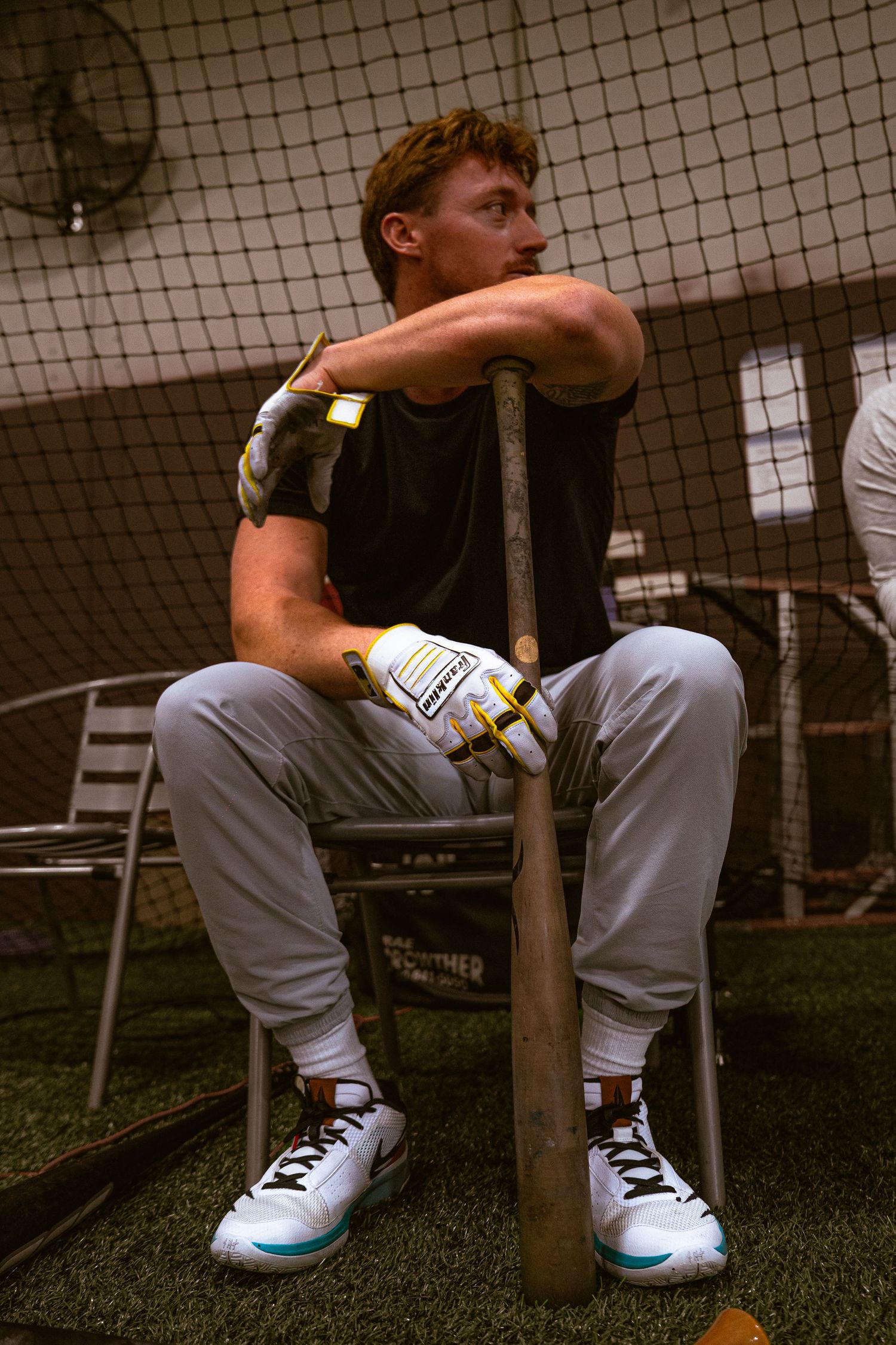 Baseball player in batting gloves sits resting on a bat inside a batting cage wearing gray pants and white cleats.
