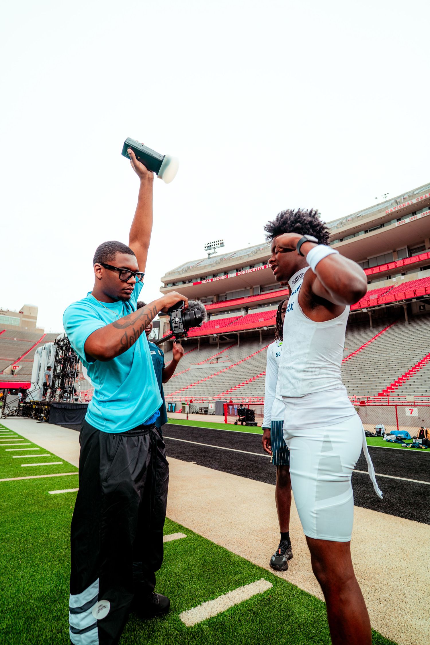 Athletic trainer assists football player on stadium sideline, holding equipment overhead during pre-game preparation.