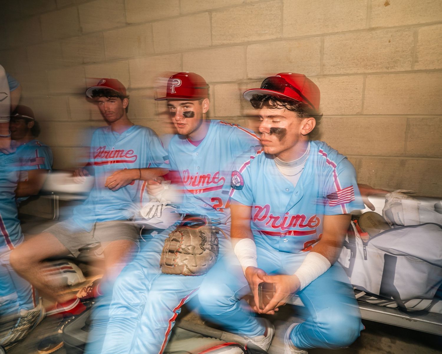 Baseball players in light blue Prime uniforms with red caps resting in a dugout during a game.