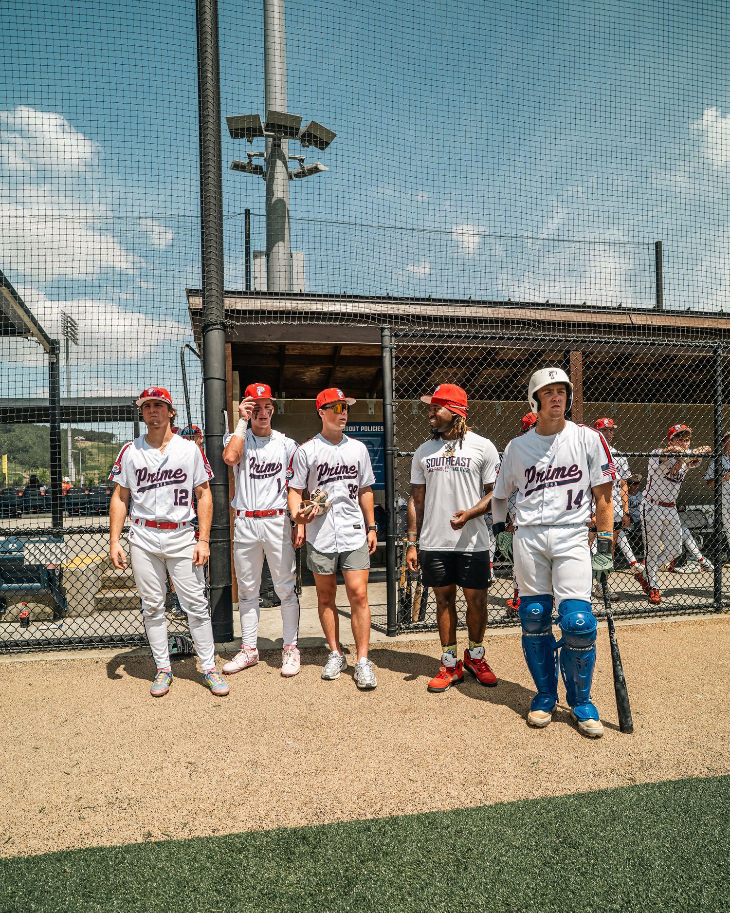Five baseball players in white Primos uniforms pose together near dugout fence on sunny game day.