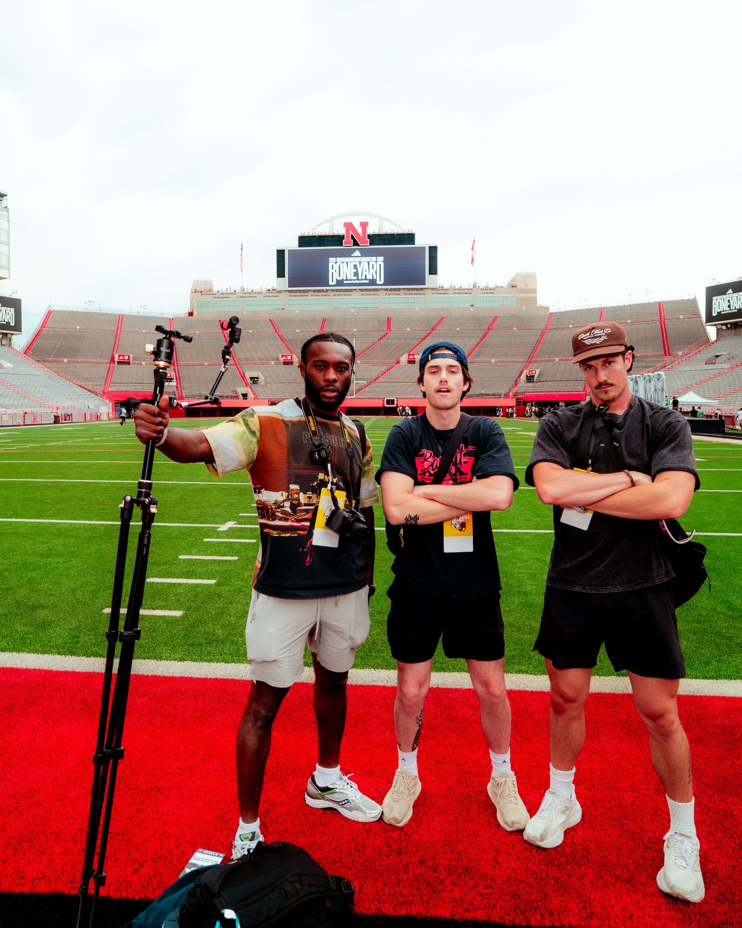 Three men pose on a football field at Nebraska Cornhuskers stadium with camera equipment.