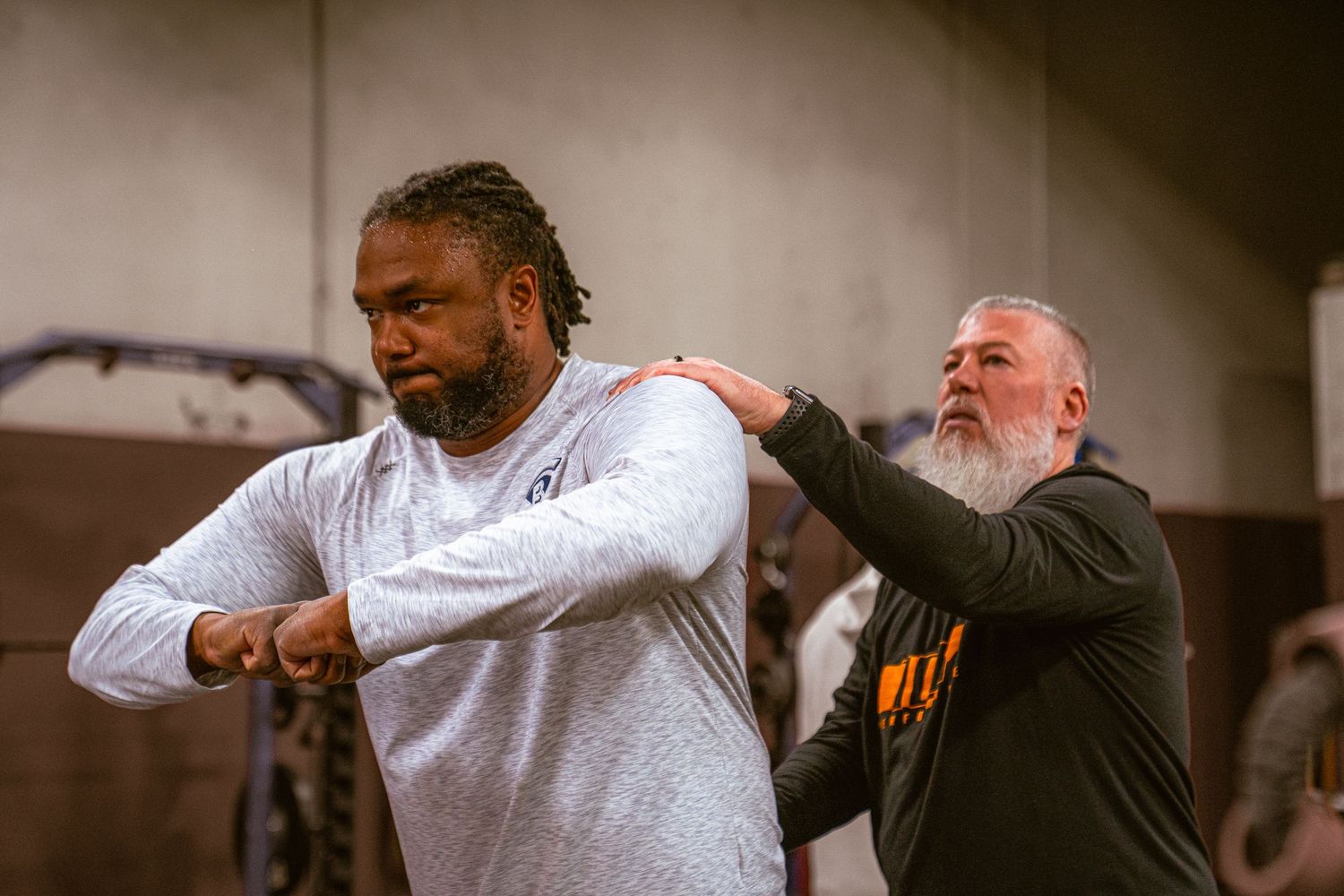 A trainer assists a large athlete with shoulder stretches during a gym training session.
