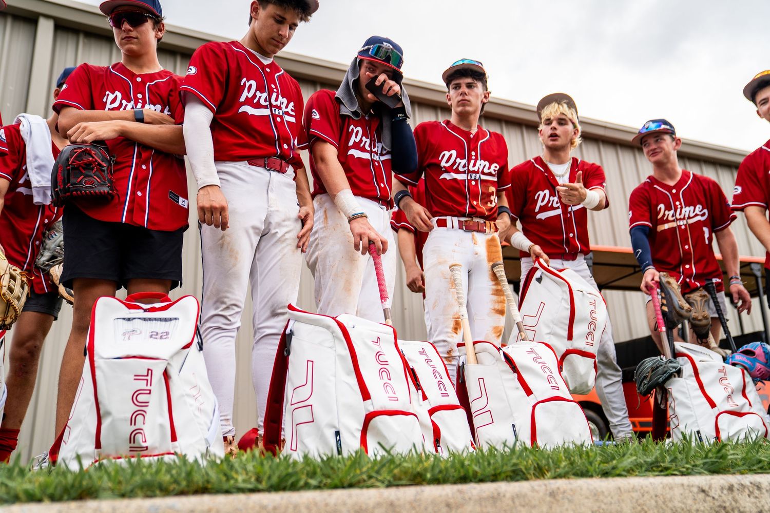 Youth baseball team in Prime uniforms standing with white equipment bags on a sunny day.