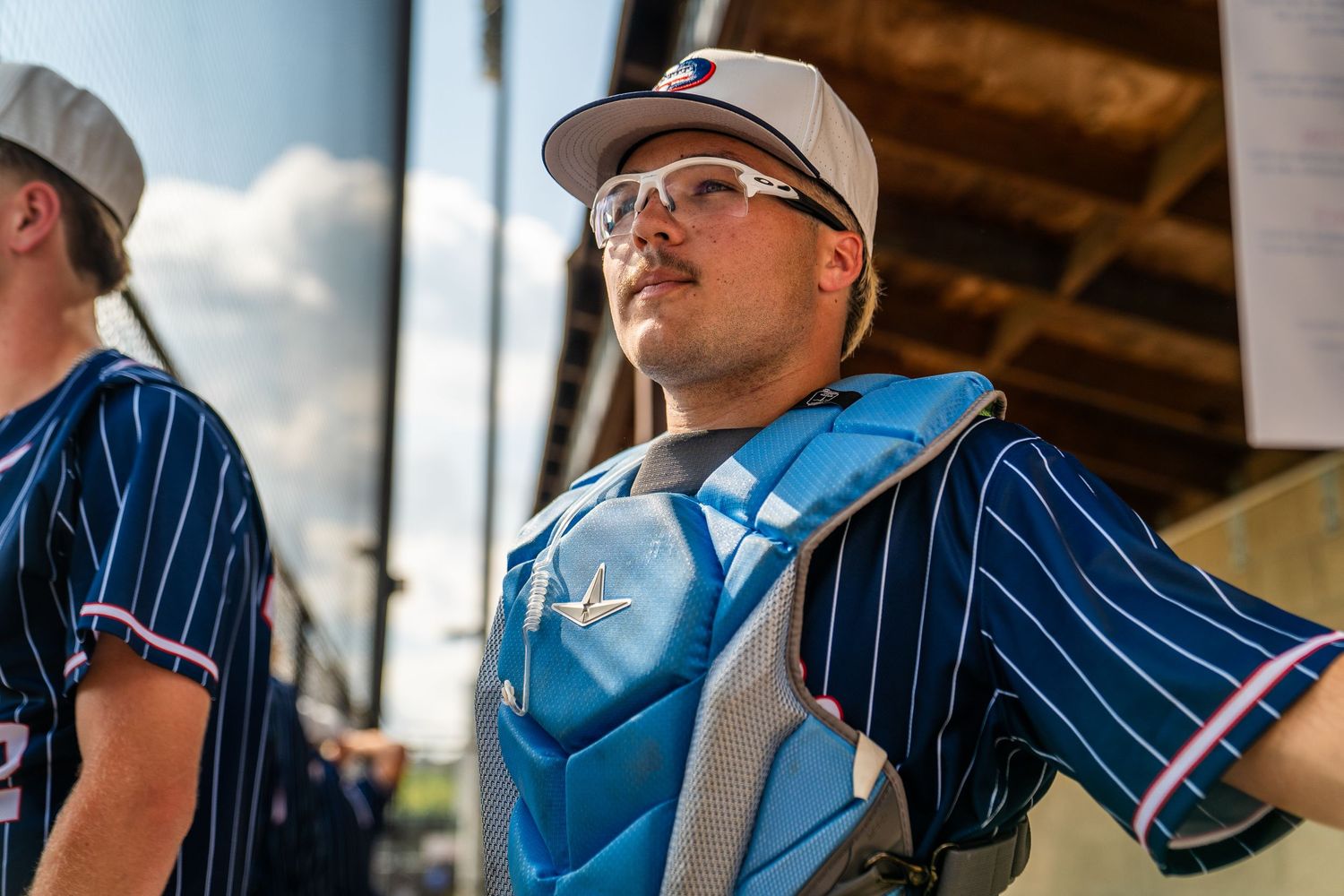 Baseball catcher in blue pinstripe jersey and chest protector wearing glasses stands in dugout during game.