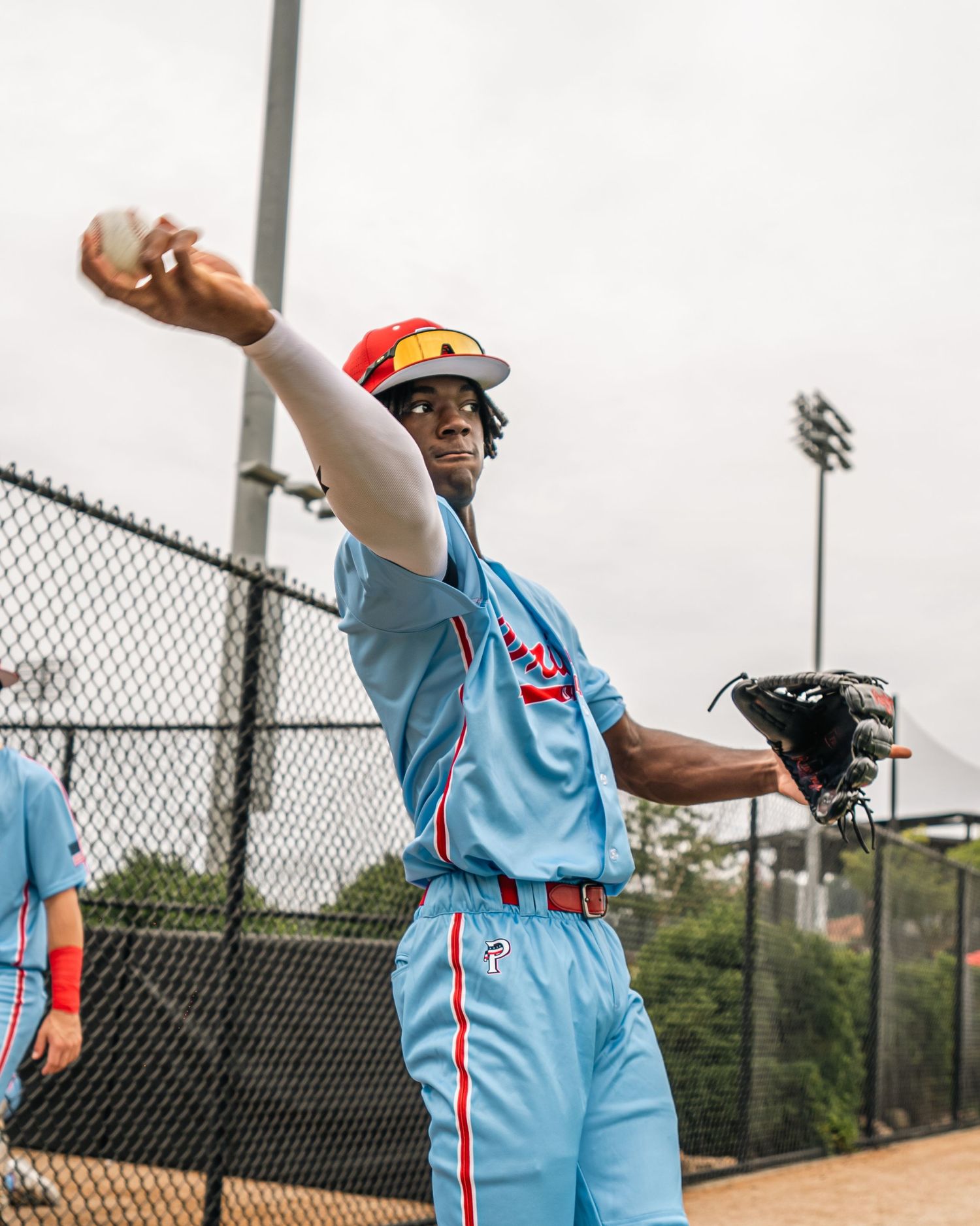 Young baseball player in light blue uniform winds up to throw a ball on an outdoor field.