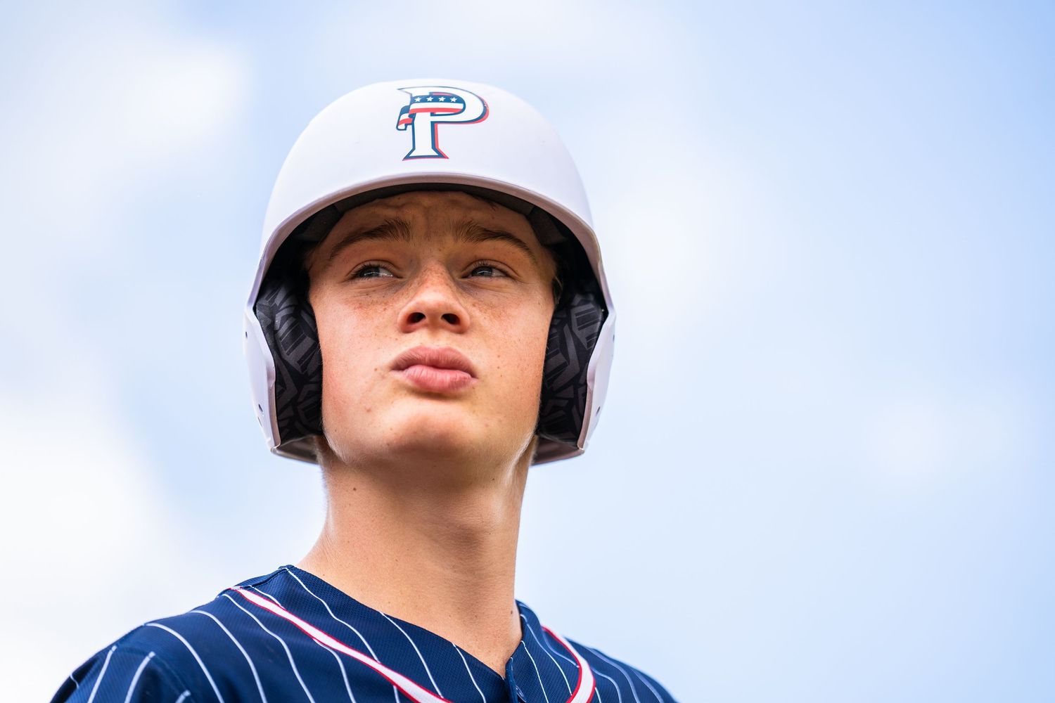 Young baseball player wearing a white helmet with P logo and navy pinstripe jersey poses outdoors.