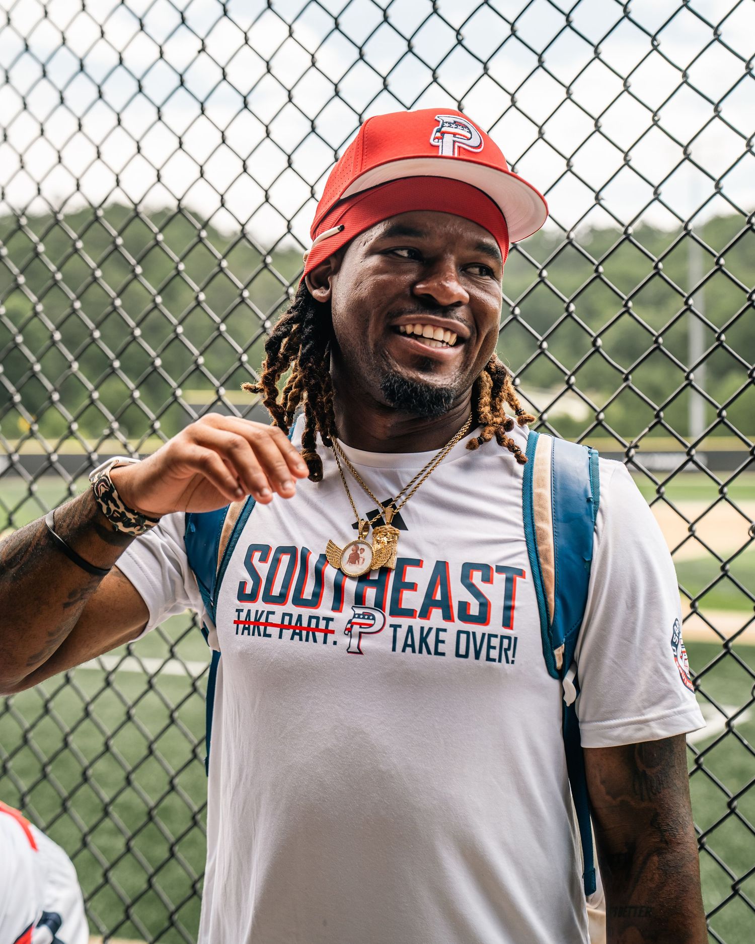 Smiling man wearing red Primos cap and Southeast Take Over shirt stands near baseball field fence.
