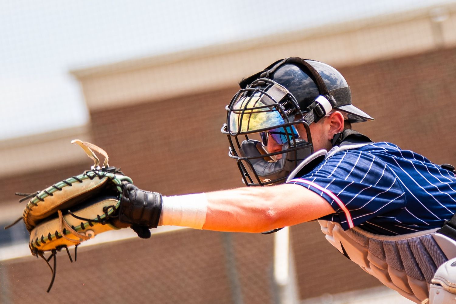 Baseball catcher in full gear reaching out with glove to catch a pitch during a sunny outdoor game.