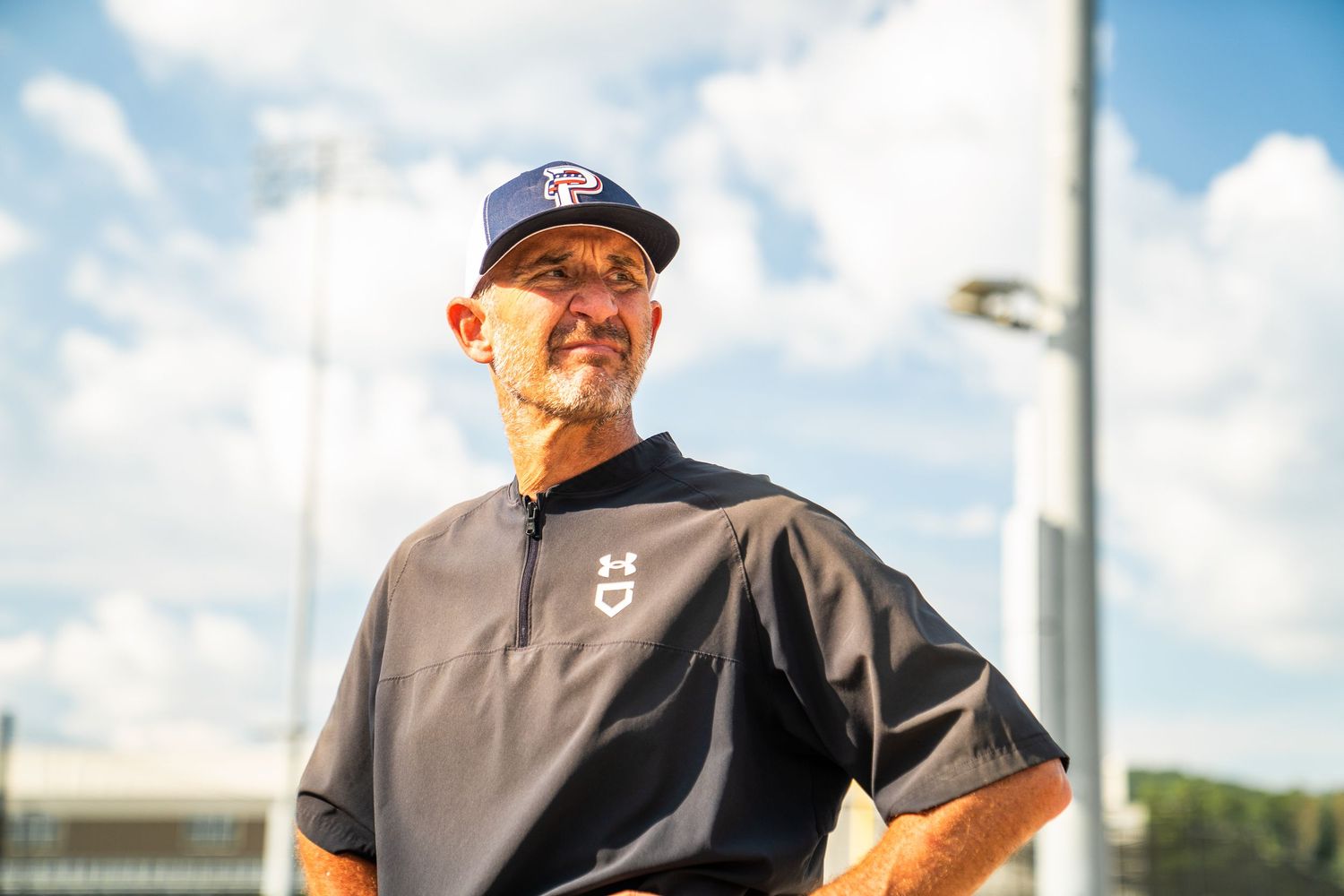 A mature baseball coach in a dark jacket and navy cap stands confidently on a sunny baseball field.