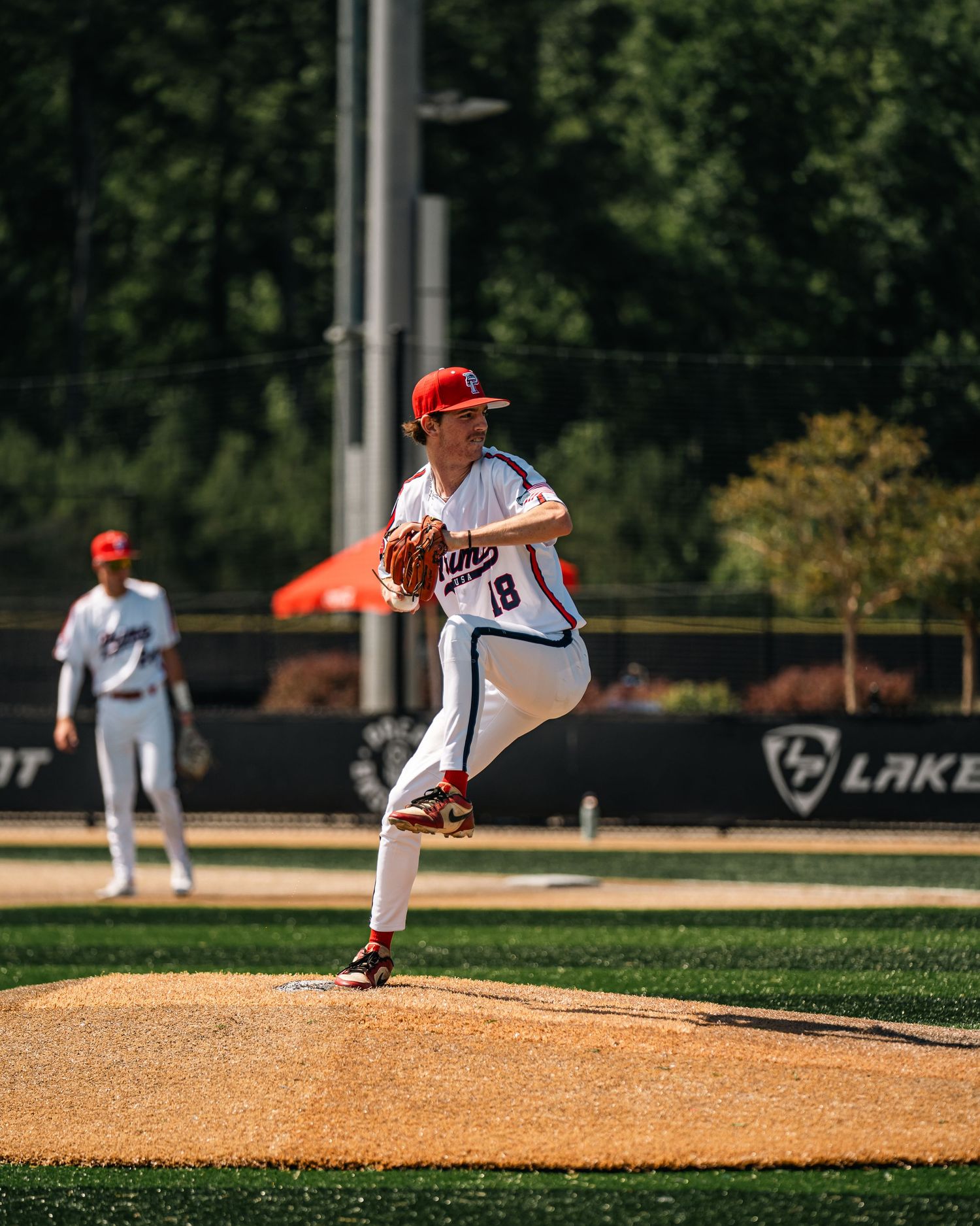 Baseball pitcher in white uniform #18 winds up on the mound during a sunny outdoor game with green trees in background.