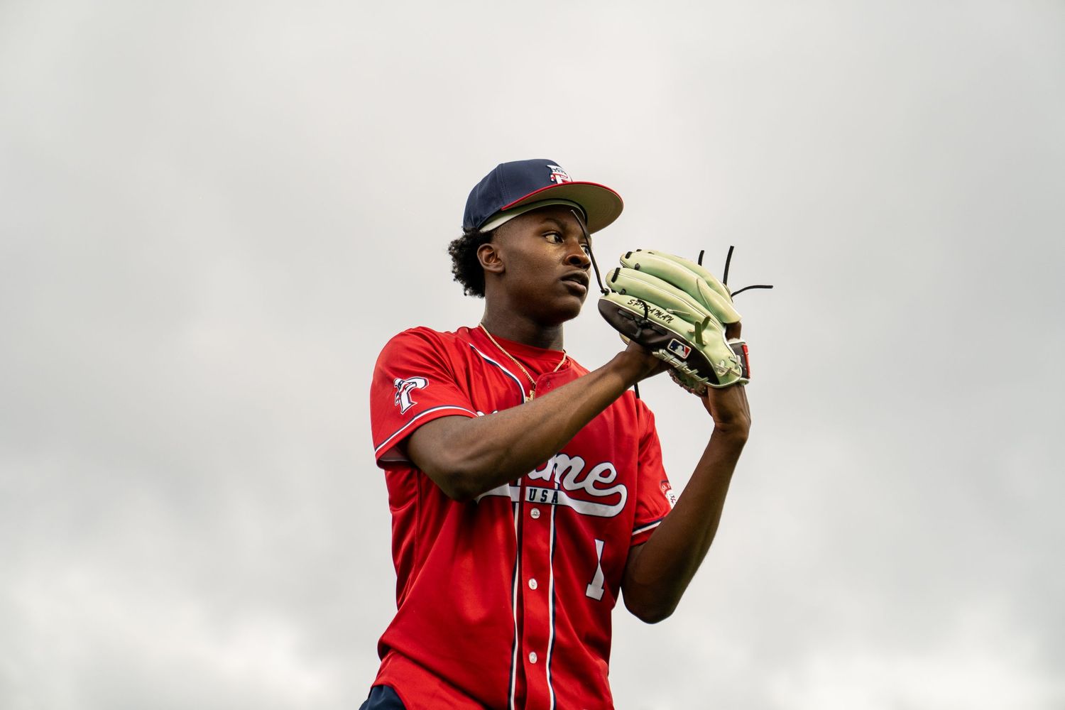 Young baseball pitcher in red uniform and navy cap holds glove ready on overcast day.