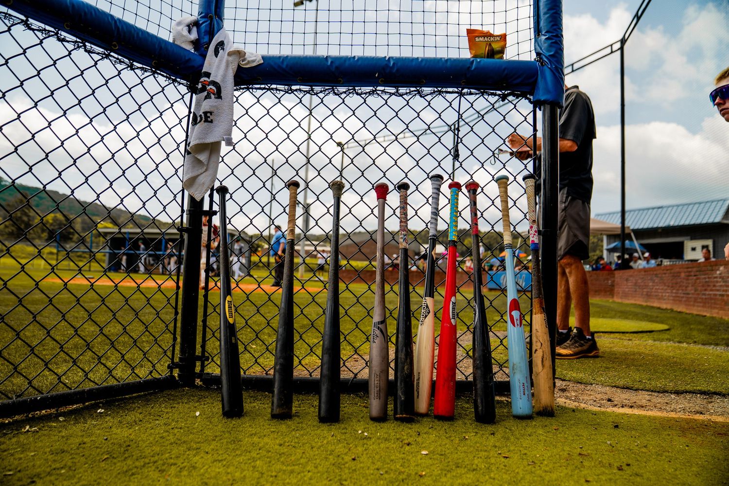 Baseball bats lined up against a chain-link fence dugout at a youth baseball field during a game day.