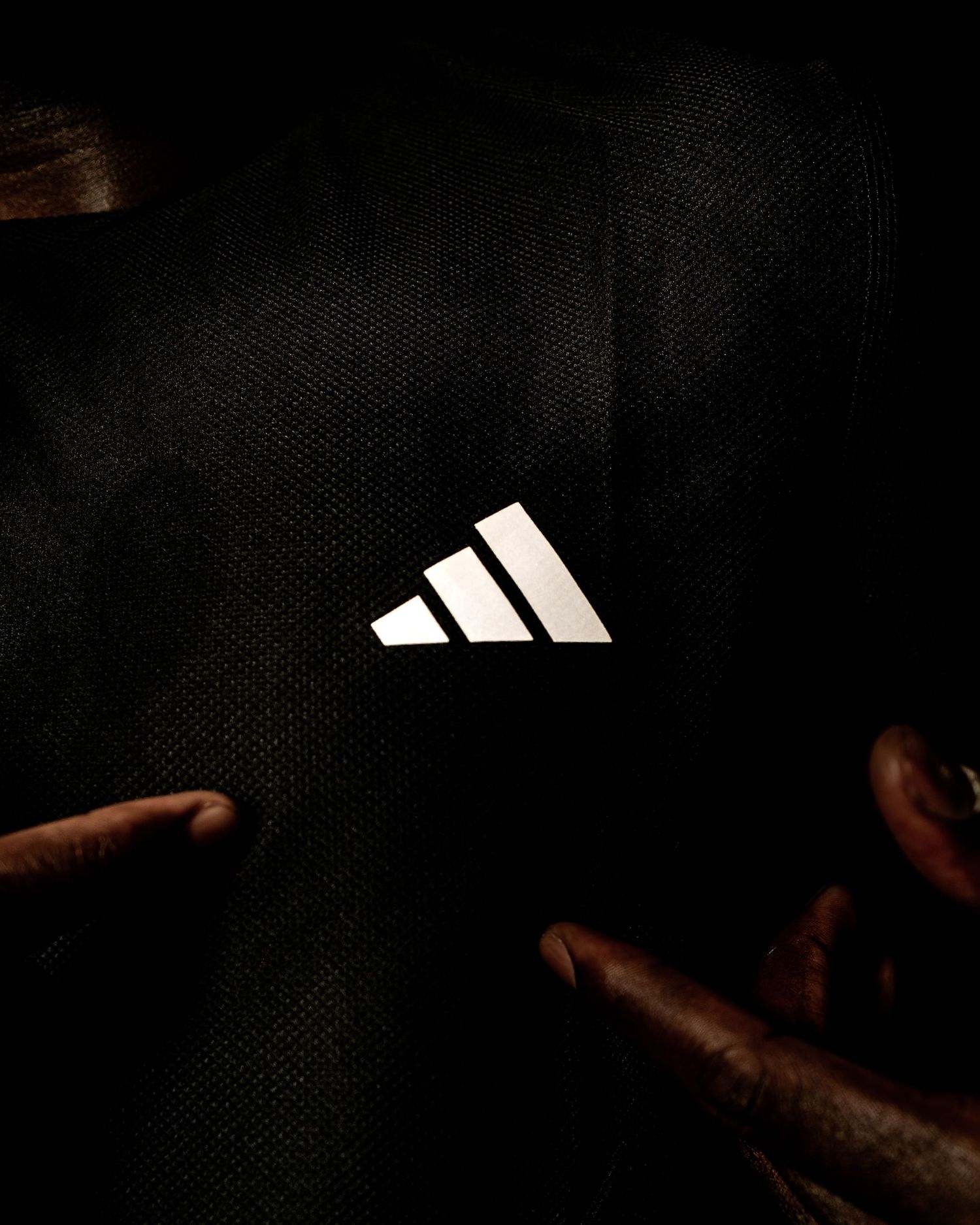 Close-up of glowing Adidas logo on dark black fabric, illuminated by fingers pointing at the reflective three-stripe emblem.