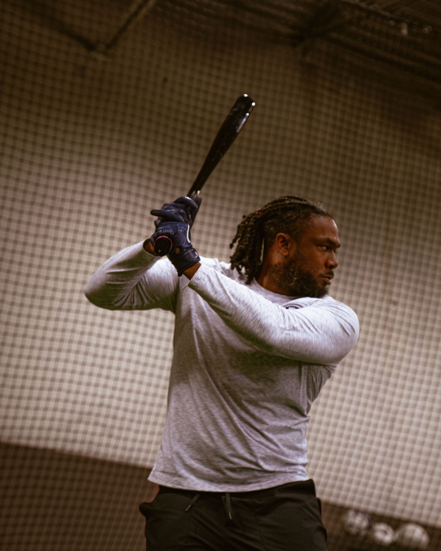 Baseball player in batting stance holding black bat in indoor batting cage wearing white long sleeve shirt.
