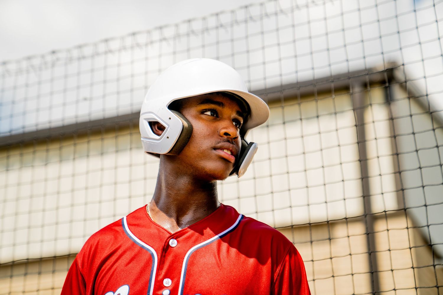 Young baseball player in red uniform and white batting helmet stands near a chain-link fence backstop.