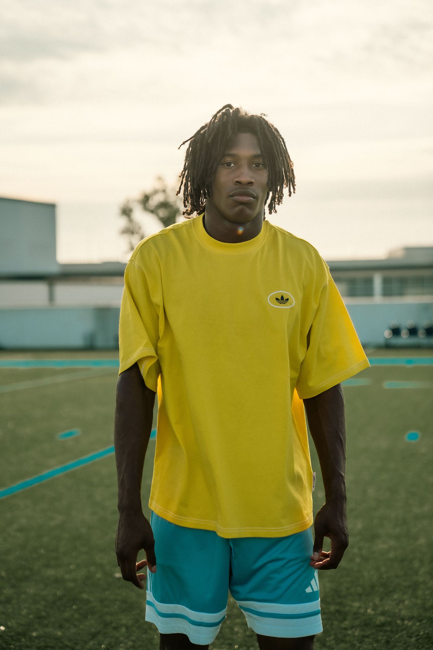Young man with dreadlocks wearing yellow Adidas t-shirt and teal shorts standing on a soccer field.