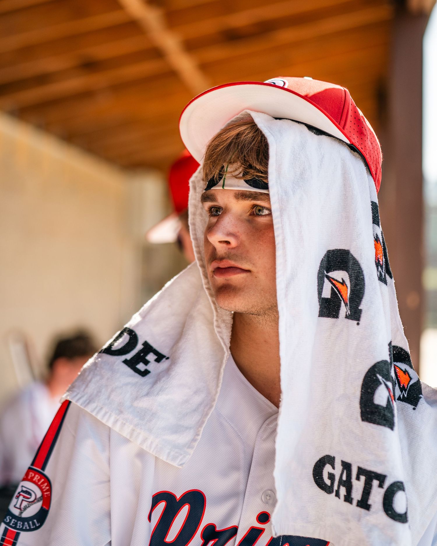 Young baseball player with towel draped over head sits in dugout wearing red and white uniform.