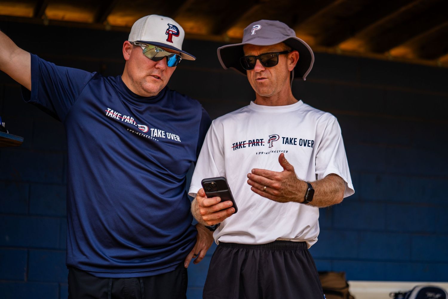 Two men in matching Take Part Take Over shirts review a phone together outdoors at a baseball facility.